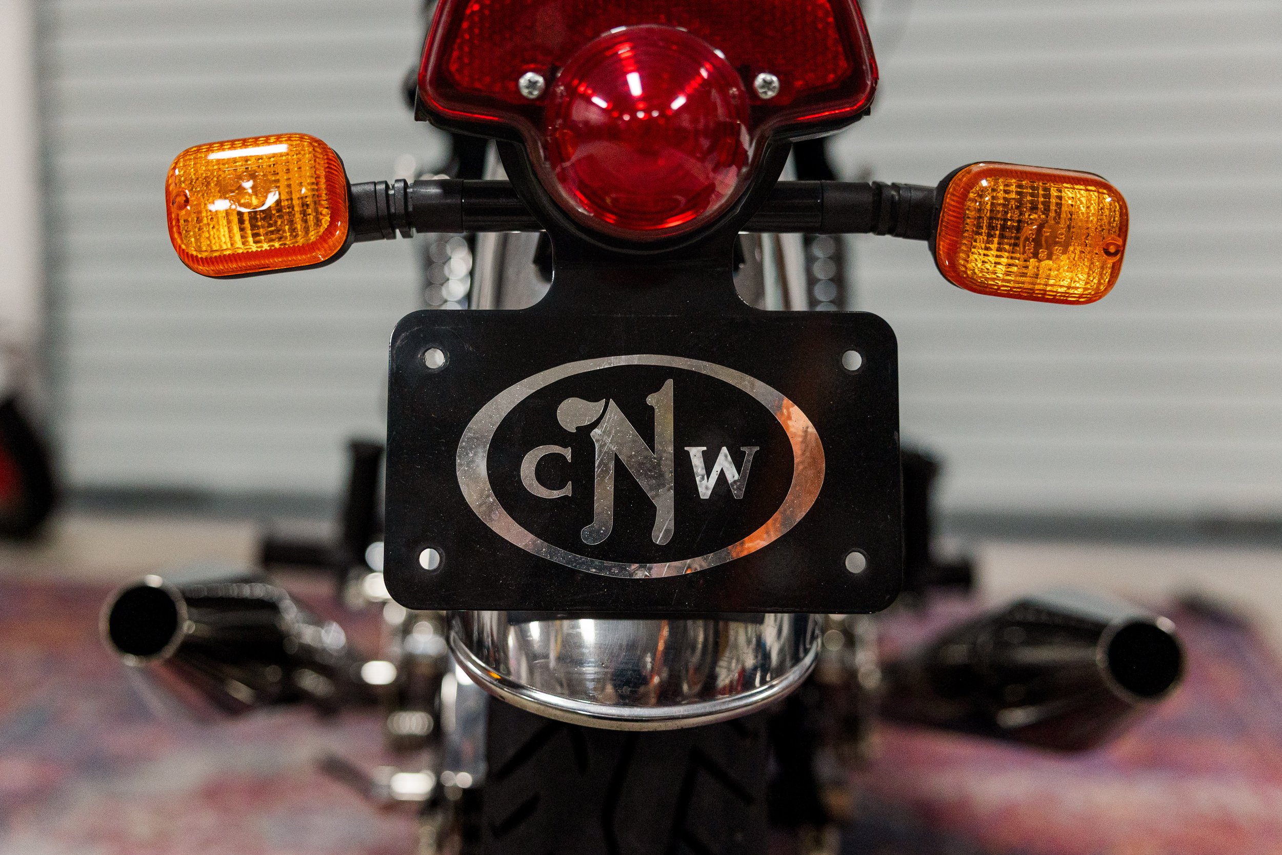 Close-up of a motorcycle's rear, showing the red tail light, orange turn signals, and a license plate with a logo. Vintage motorcycle detail at Birmingham Vintage Cycle, a vintage motorcycle restoration shop in Birmingham Alabama