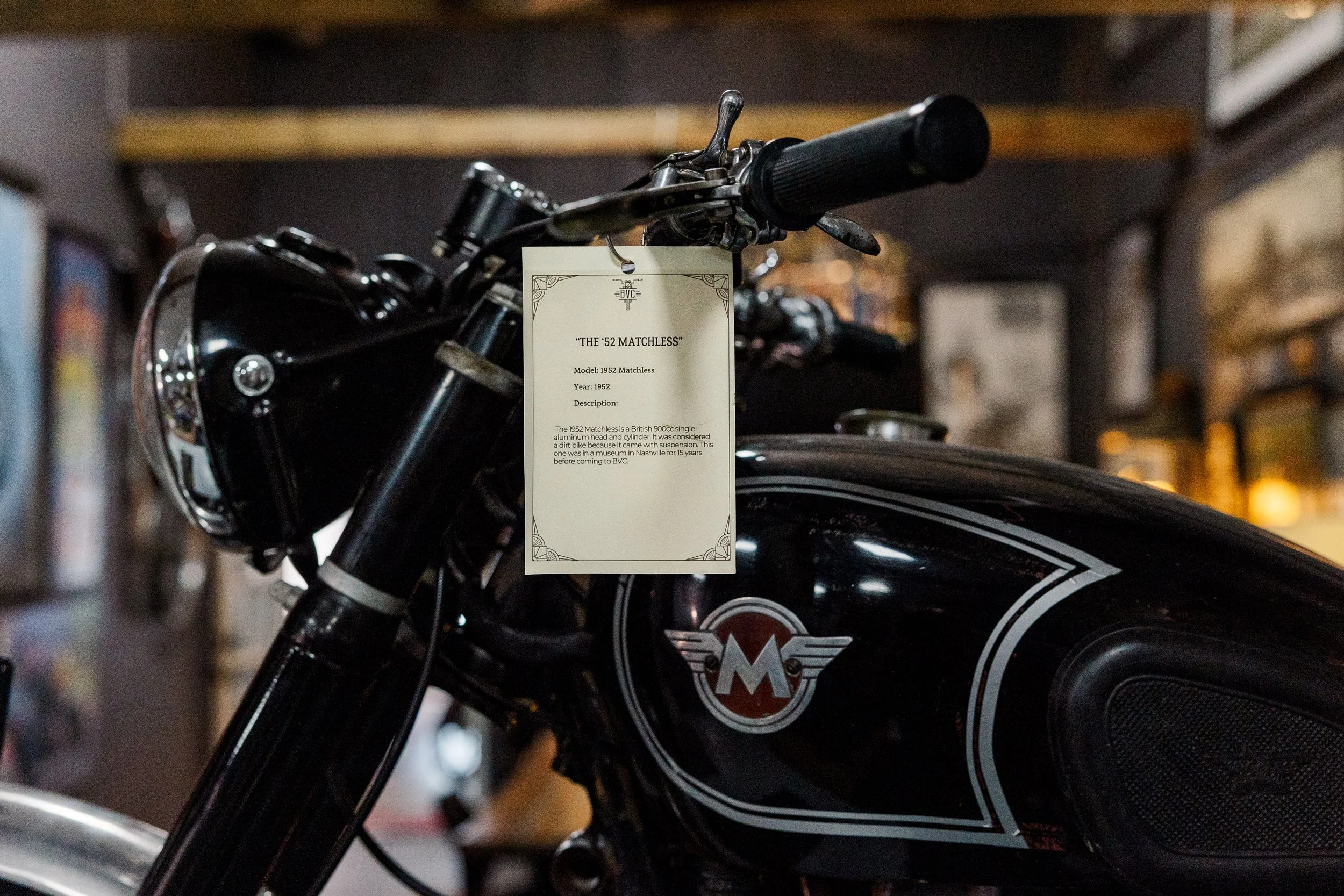 A vintage black motorcycle with a white and red badge on the side, on display in a museum. There is a label in front of it describing the model as a 1952 Matchless, a British 500cc single aluminum head and cylinder dirt bike with suspension, exhibite