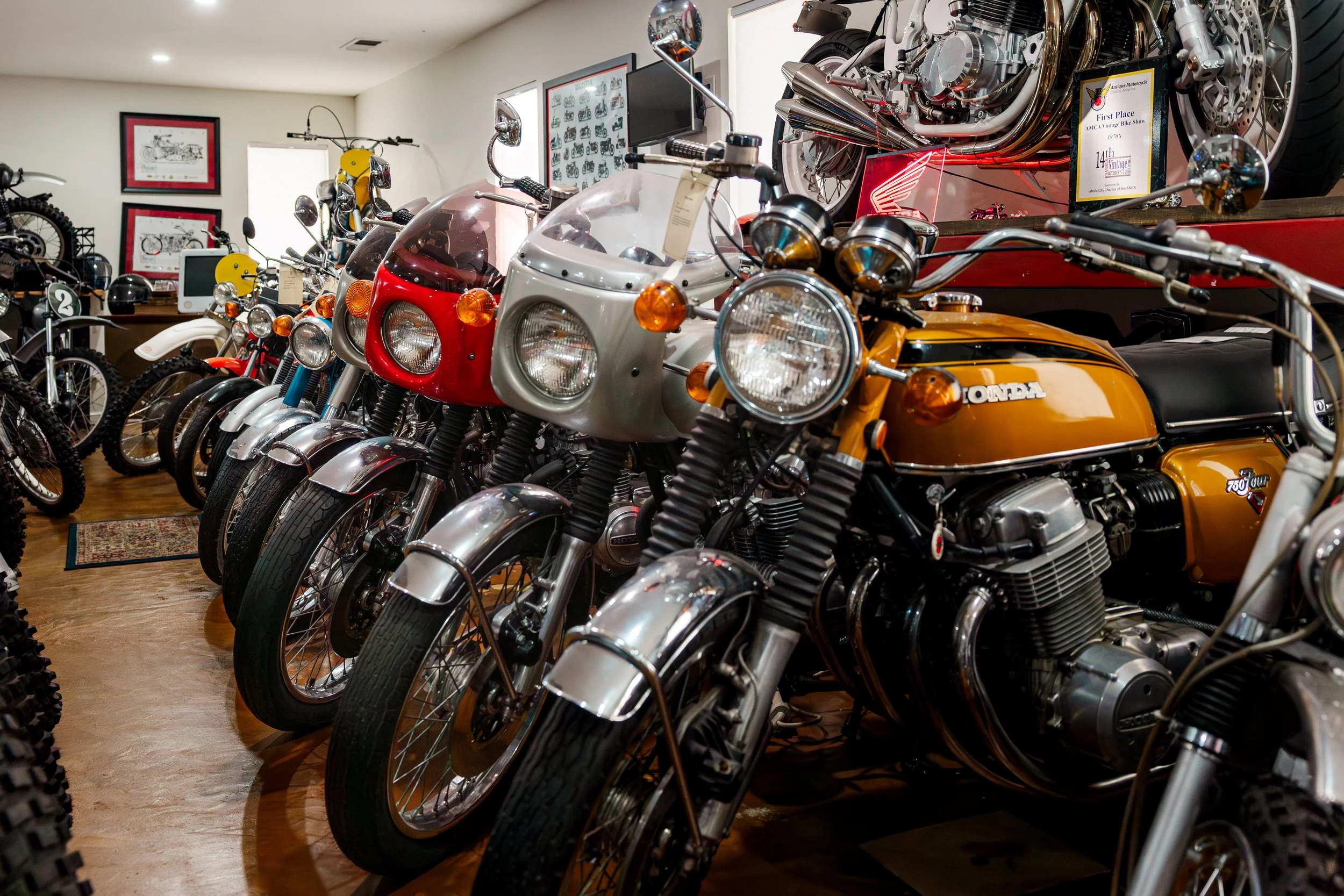 A collection of vintage motorcycles displayed inside a museum, including a Honda with orange and black detailing and a white and red motorcycle, with framed pictures and documentation on the walls. Vintage motorcycle collection on display at BVC