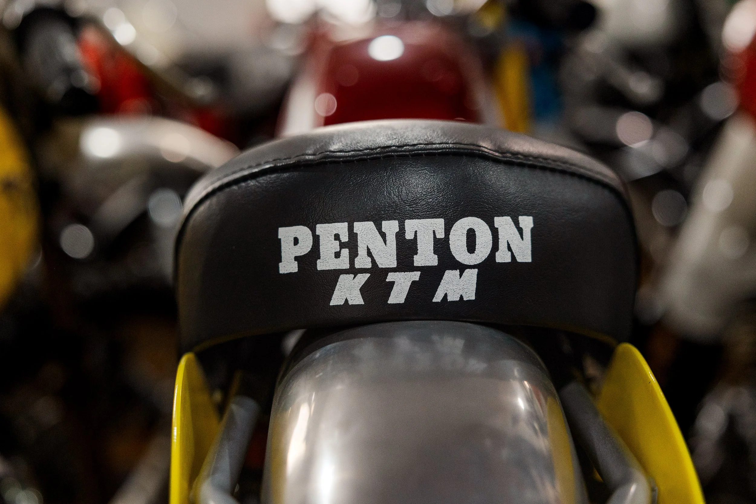 A close-up view of a luggage cart with a black leather pad that has the words 'PENTON KTM' printed on it, surrounded by other luggage in various colors and shapes, with a focus on the pad. Vintage motorcycle detail at Birmingham Vintage Cycle, 