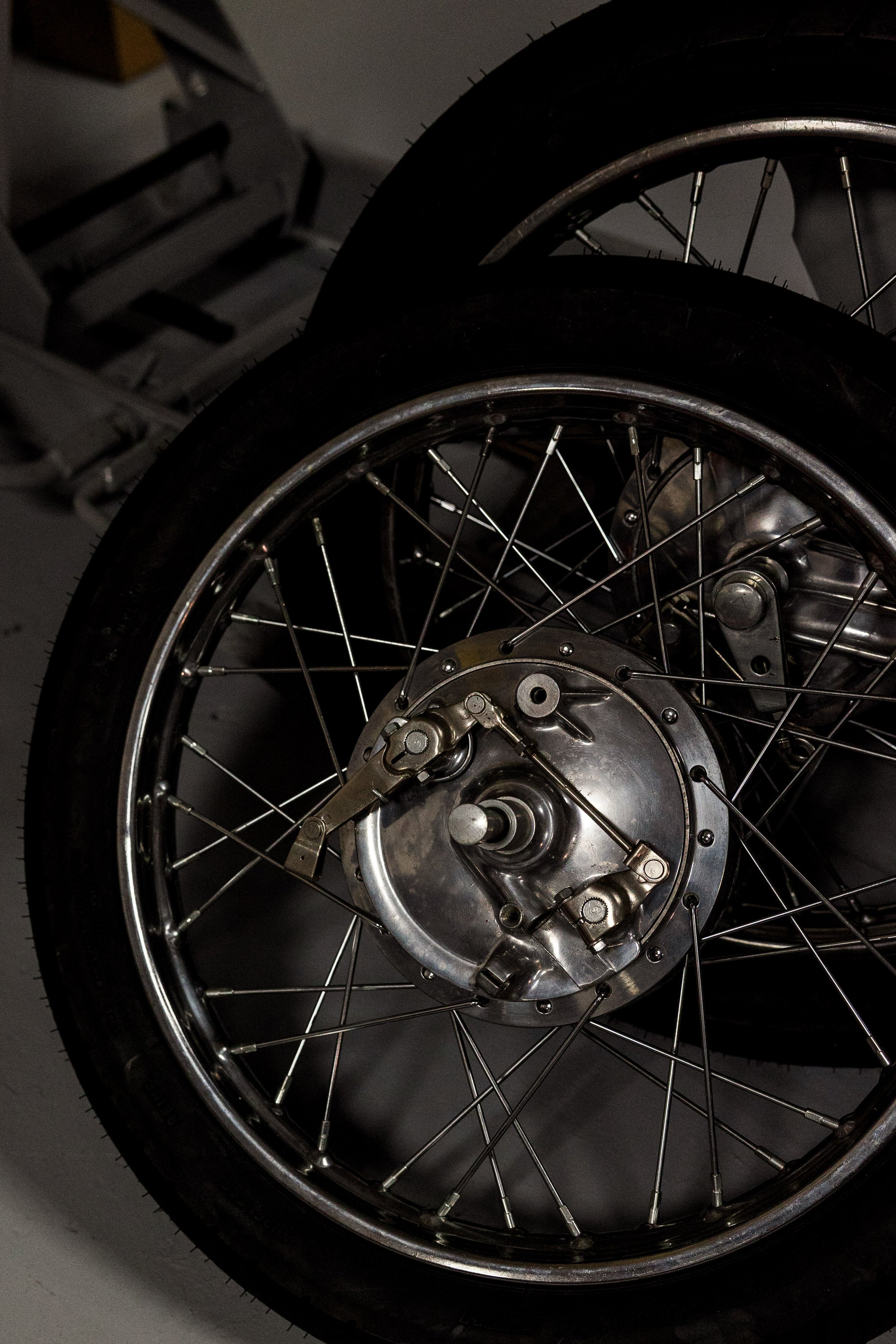 Close-up of a motorcycle wheel with black tire, silver spokes, and metallic hub, set against a dark background. Vintage motorcycle collection on display at Birmingham Vintage Cycle in Birmingham Alabama