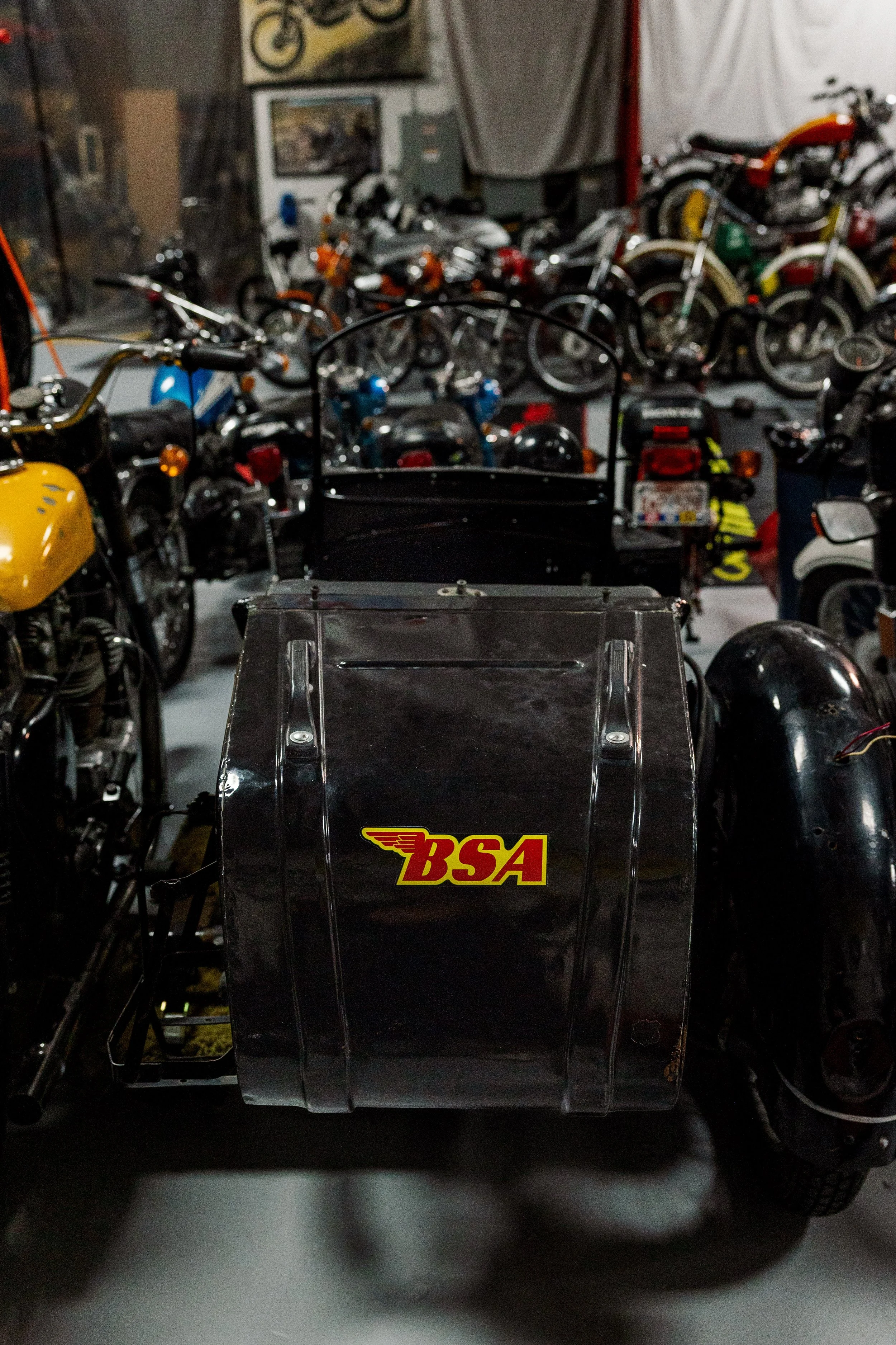 View of a collection of vintage motorcycles in a dimly lit garage or showroom, with a black IVG racing cart in the foreground and various motorcycle models on display background. Vintage motorcycle collection on display at Birmingham Vintage Cycle