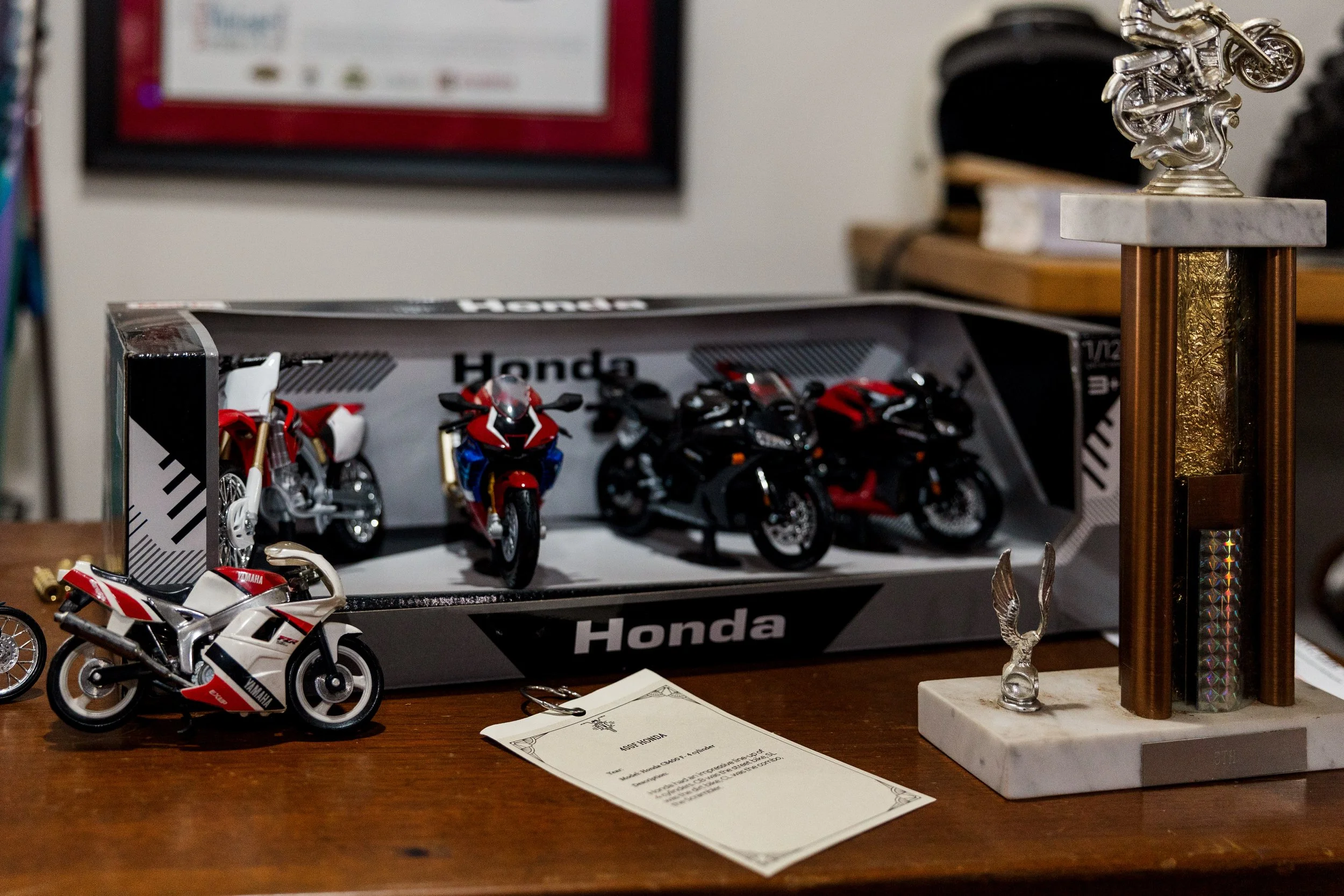 Model motorcycles and a trophy on a wooden table, with a box of miniature Honda motorcycles in the background and a framed certificate or award on the wall. Inside Birmingham Vintage Cycle, a vintage motorcycle shop and community space in Birmingham 