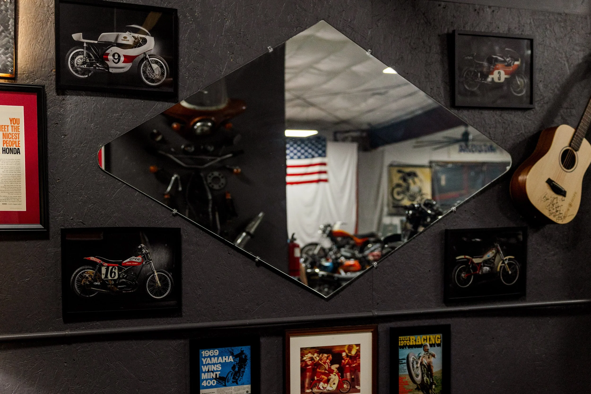 A wall decorated with framed pictures of racing motorcycles, a guitar, and various posters, along with part of a mirror reflecting an American flag and other objects.Inside Birmingham Vintage Cycle, a vintage motorcycle shop and community space