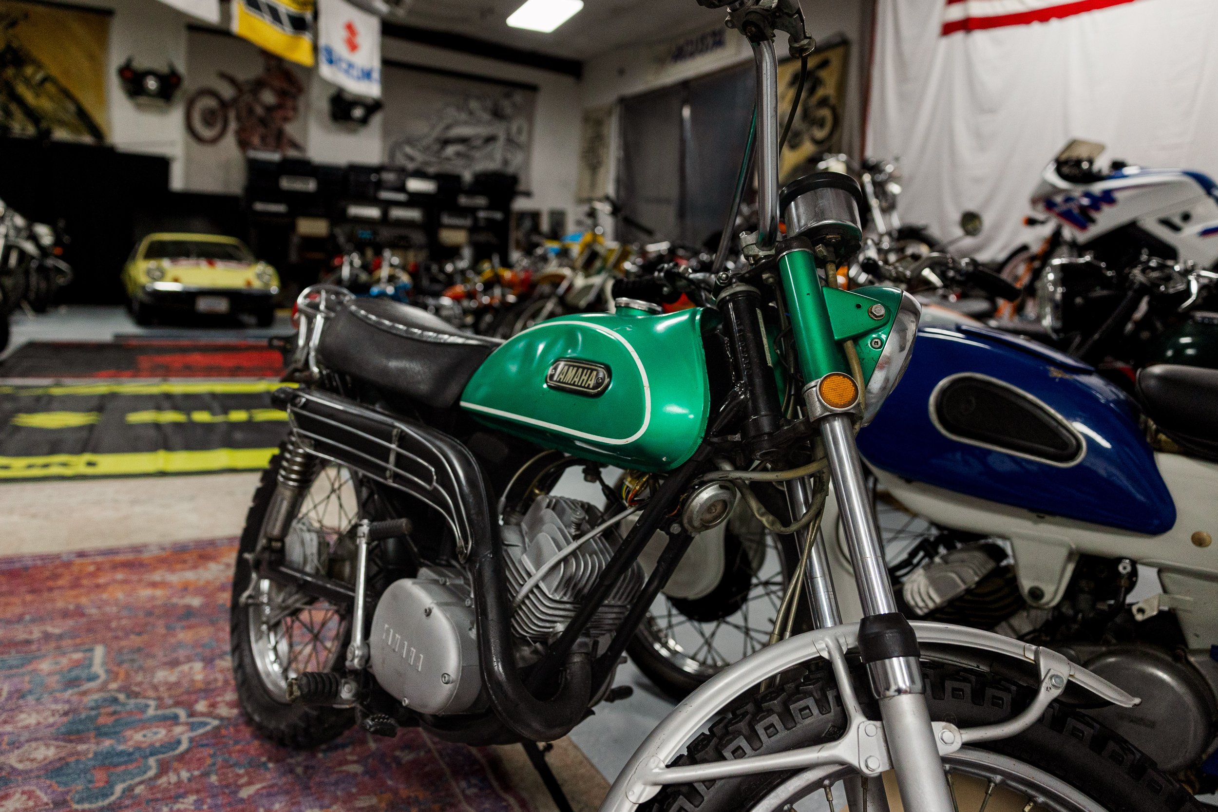 A vintage green Yamaha motorcycle in foreground, with several other motorcycles and a pile of model cars in the background inside a garage, decorated with flags and banners. Vintage motorcycle detail at Birmingham Vintage Cycle, a vintage motorcycle 