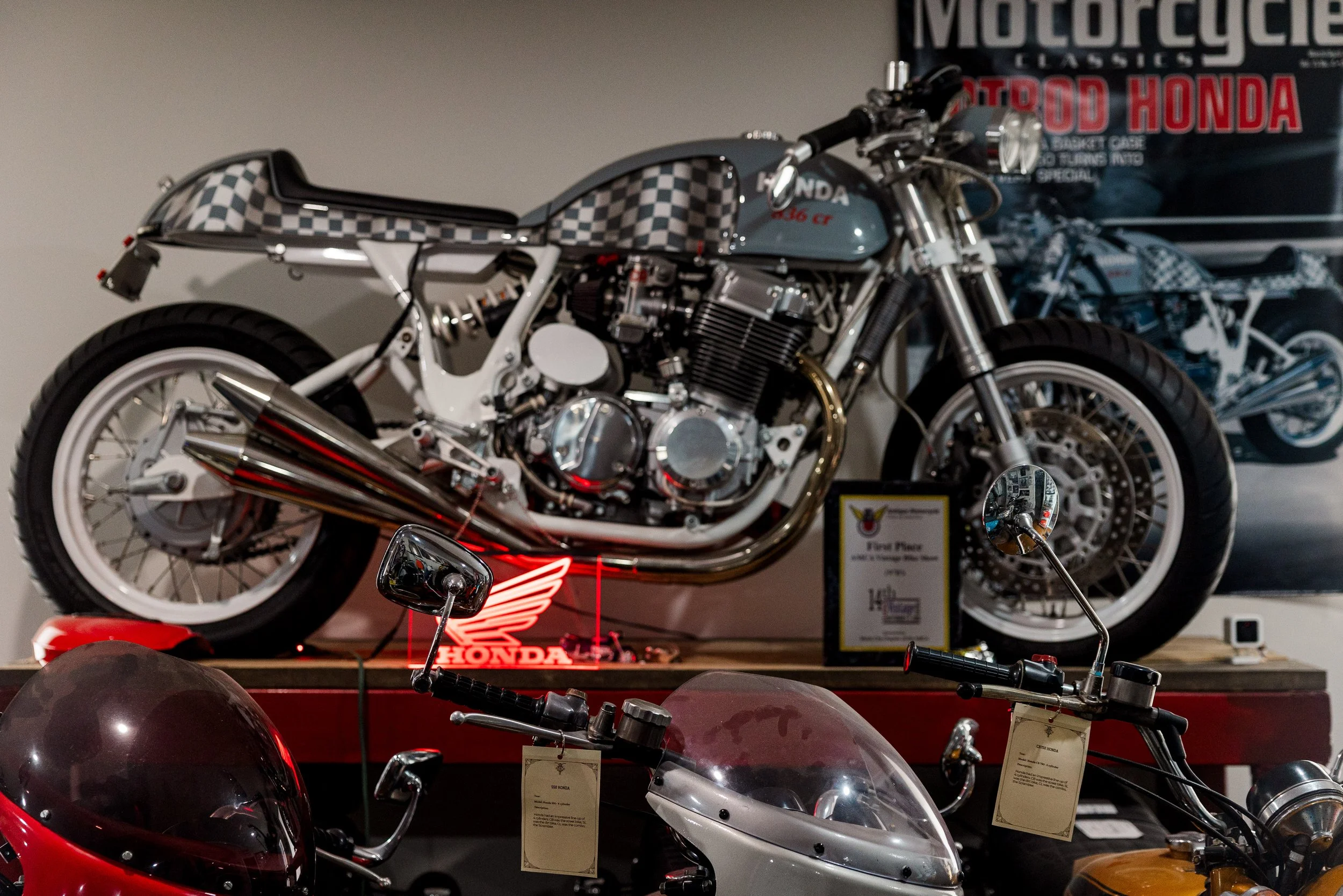 A motorcycle with a gray gas tank and checkered pattern on the seat and rear section on display at a motorcycle exhibit, with vintage bikes and Honda signage in the background. Restored vintage motorcycle in the Birmingham Vintage Cycle collection in