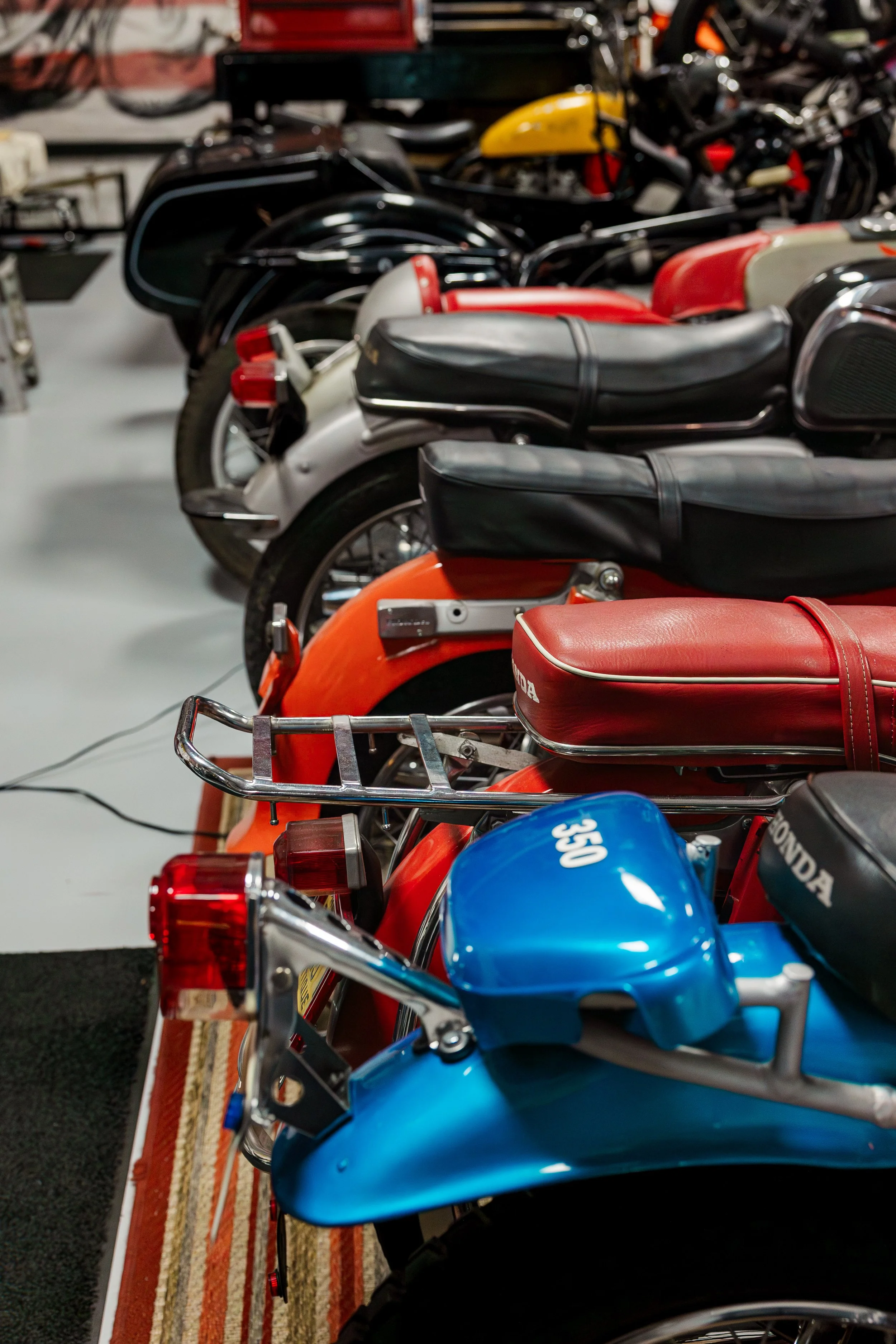 Multiple vintage Honda motorcycles in various colors including red, black, yellow, blue, and orange, parked indoors on a striped rug. Vintage motorcycle collection on display at Birmingham Vintage Cycle in Birmingham Alabama