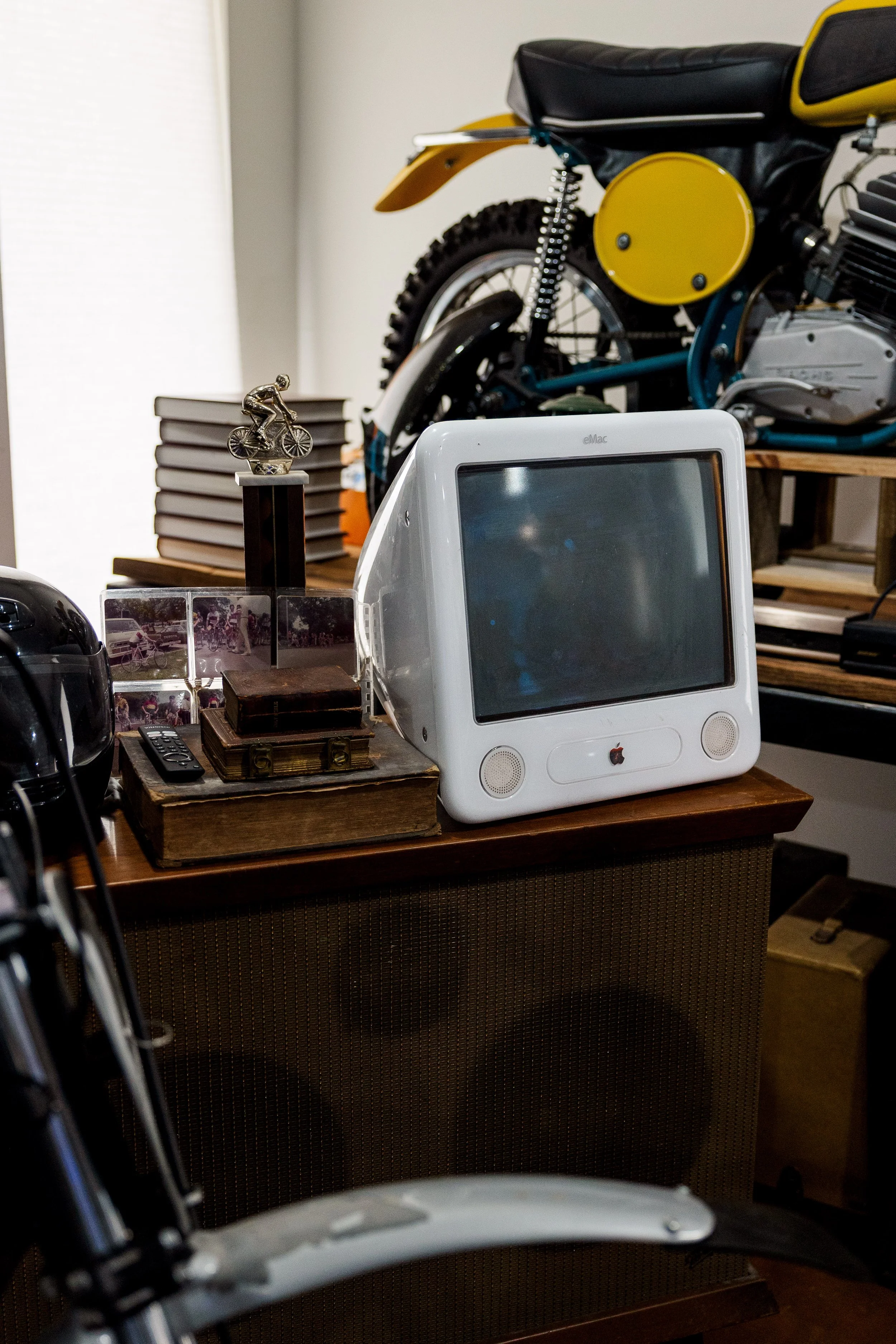 A cluttered desk with an Apple iMac G3 computer, a vintage TV remote, a miniature cyclist trophy, a stack of photographs, and a wooden box, all set on a wooden surface with a vintage motorcycle behind it. Inside Birmingham Vintage Cycle, a vintage mo