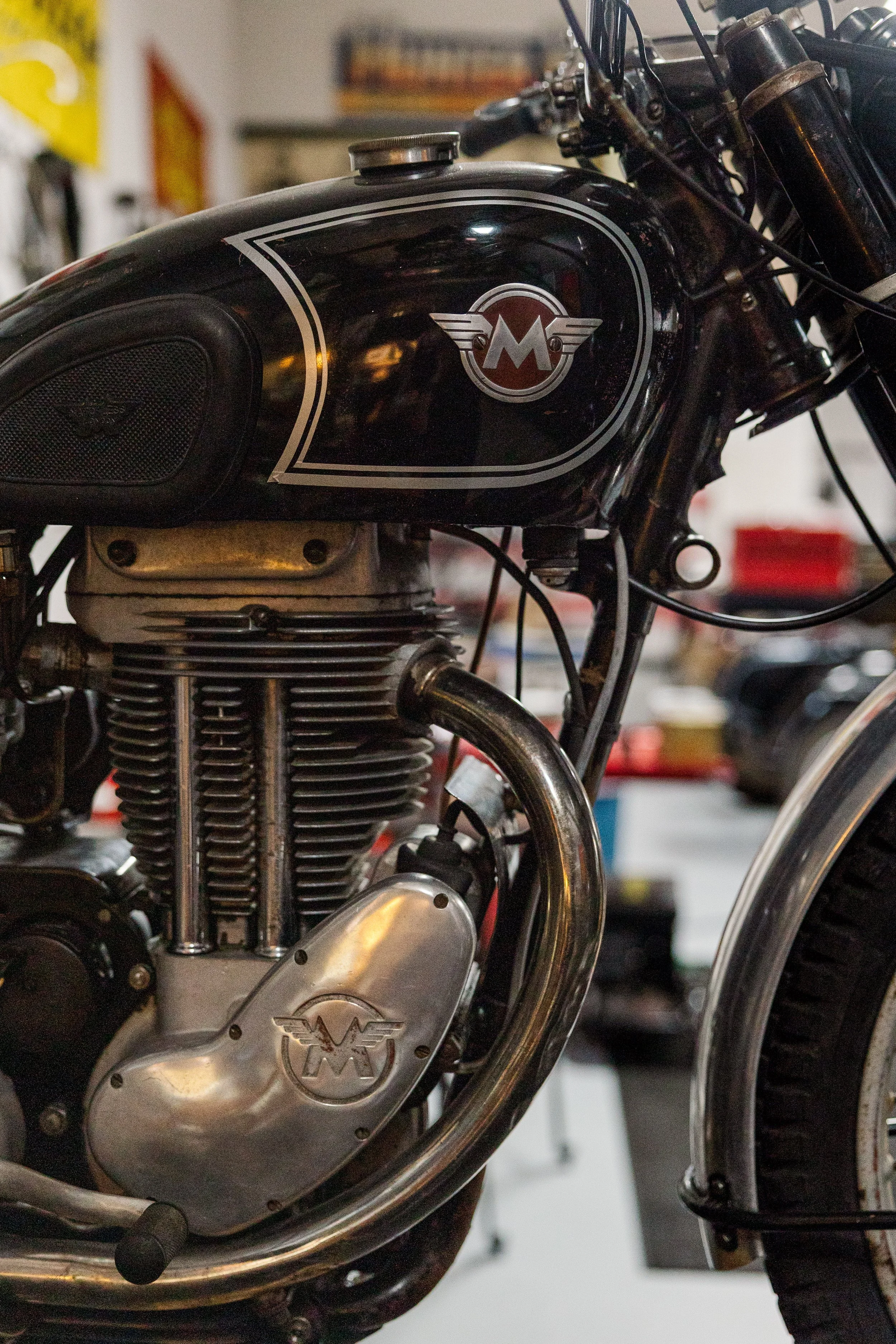 Close-up of a black vintage motorcycle gas tank with a silver emblem featuring a winged monogram, showing some mechanical parts and the engine below. Vintage motorcycle detail at Birmingham Vintage Cycle, a vintage motorcycle restoration shop in Birm