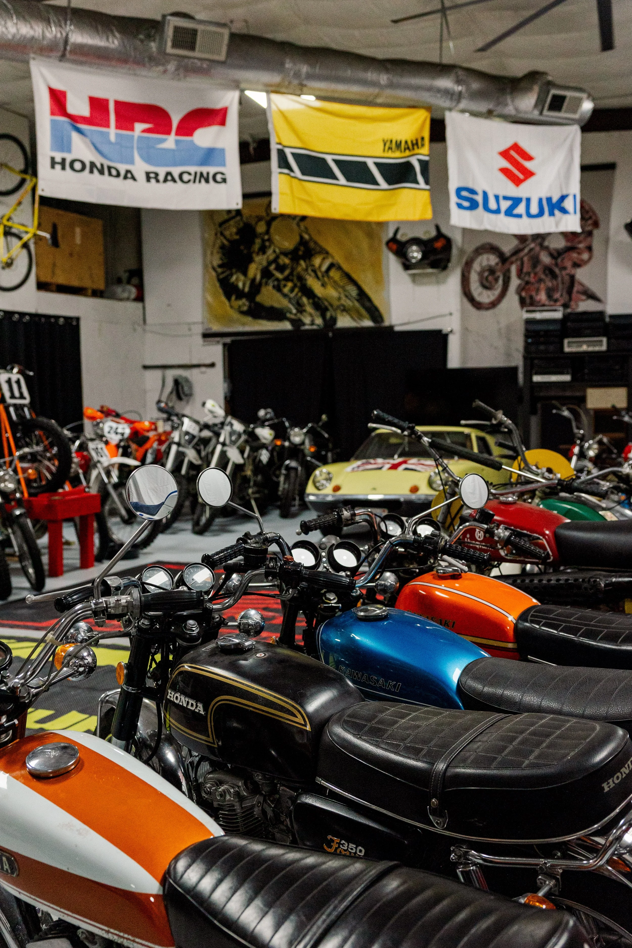 A garage with vintage motorcycles, cars, and banners from Honda Racing, Yamaha, and Suzuki hanging from the ceiling. The garage is decorated with motorcycle-themed art and memorabilia. Vintage motorcycle collection on display at BVC