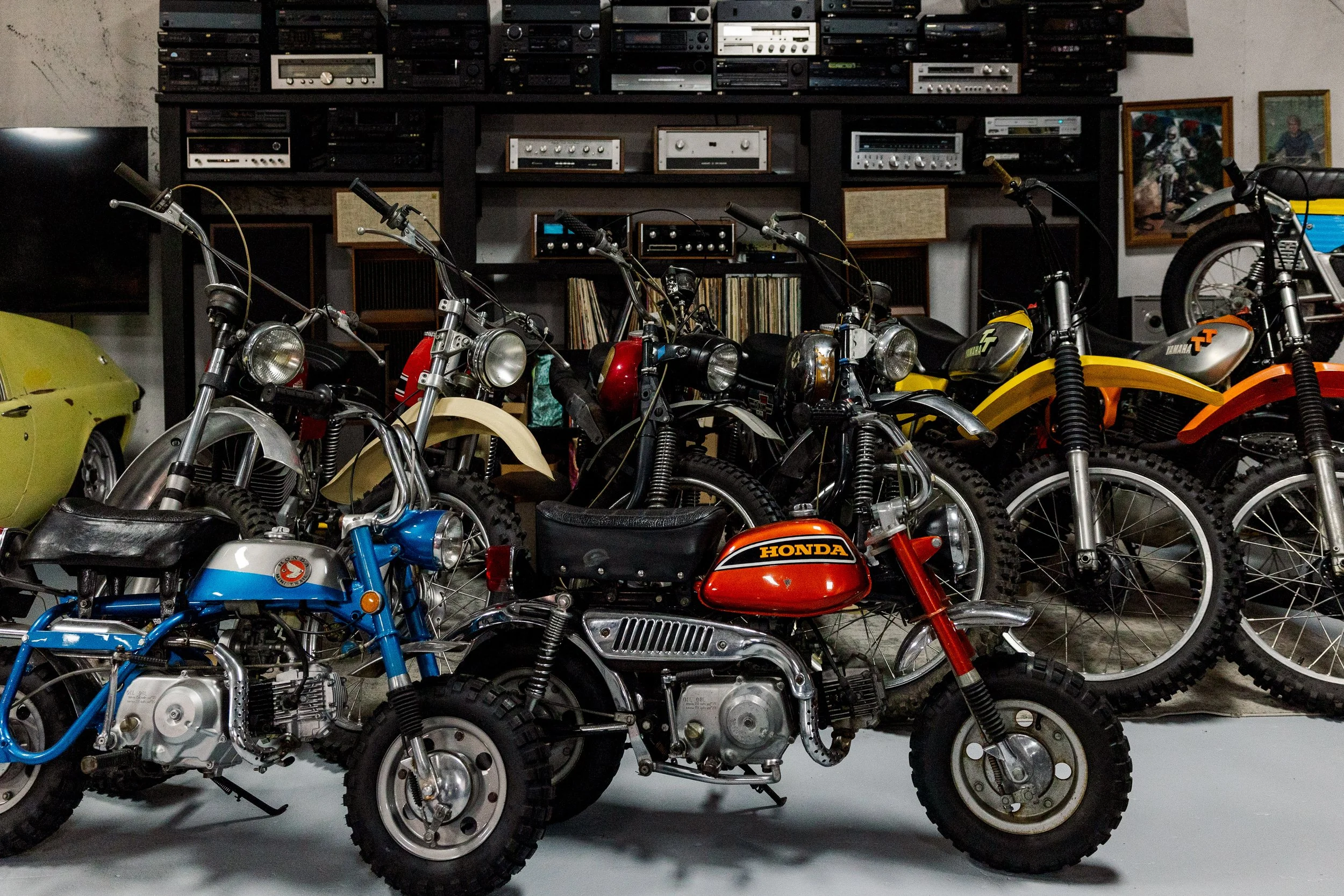 A collection of vintage motorcycles, including Honda and Yamaha models, parked indoors with a large wall of audio equipment and vinyl records in the background. Vintage motorcycle collection on display at Birmingham Vintage Cycle in Birmingham