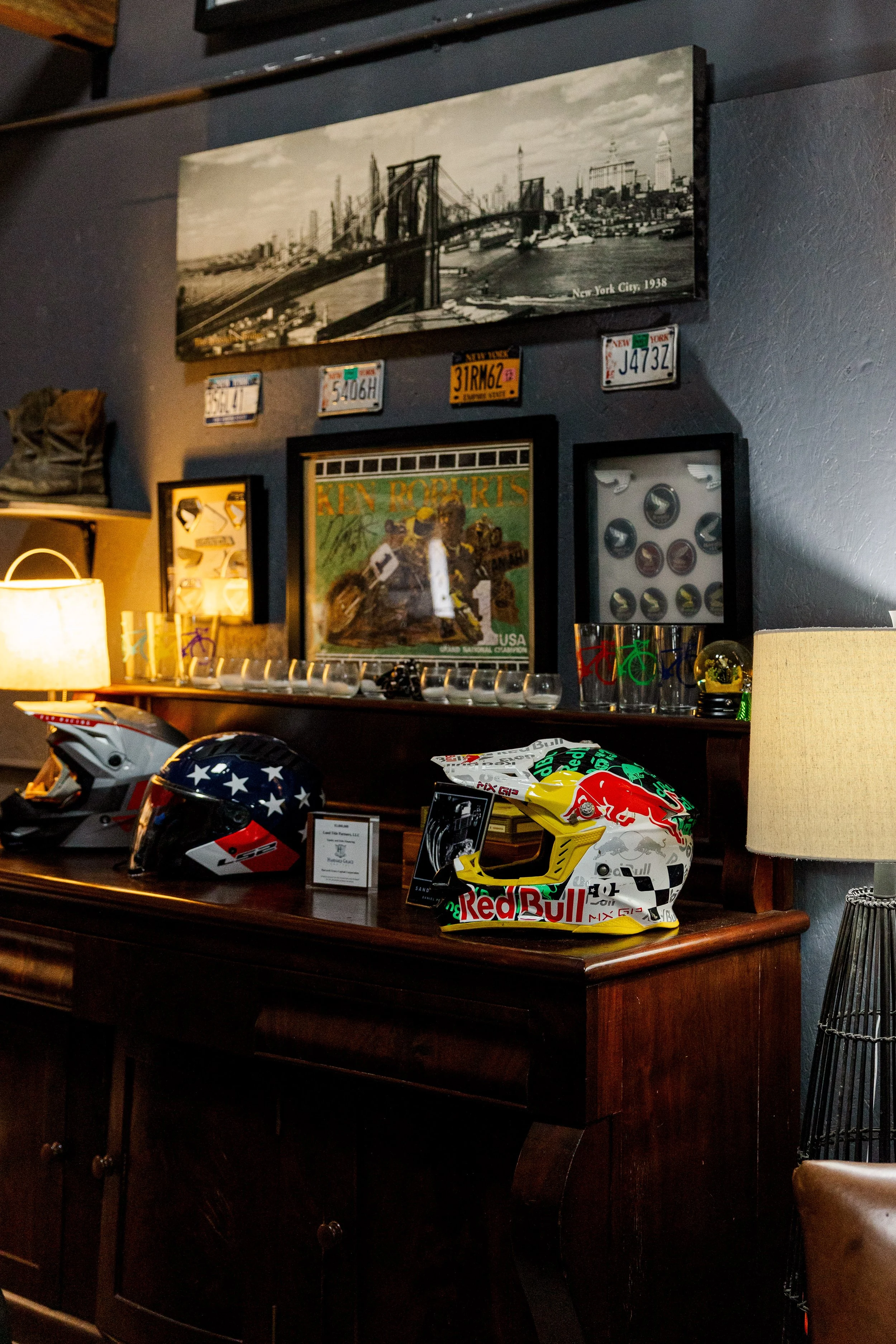 Display of several motorcycle helmets on a dark wooden cabinet, including a Red Bull racing helmet in the foreground with a white, yellow, black, and red design. Behind the helmets are framed pictures, including helmets with logos and a photo of a mo