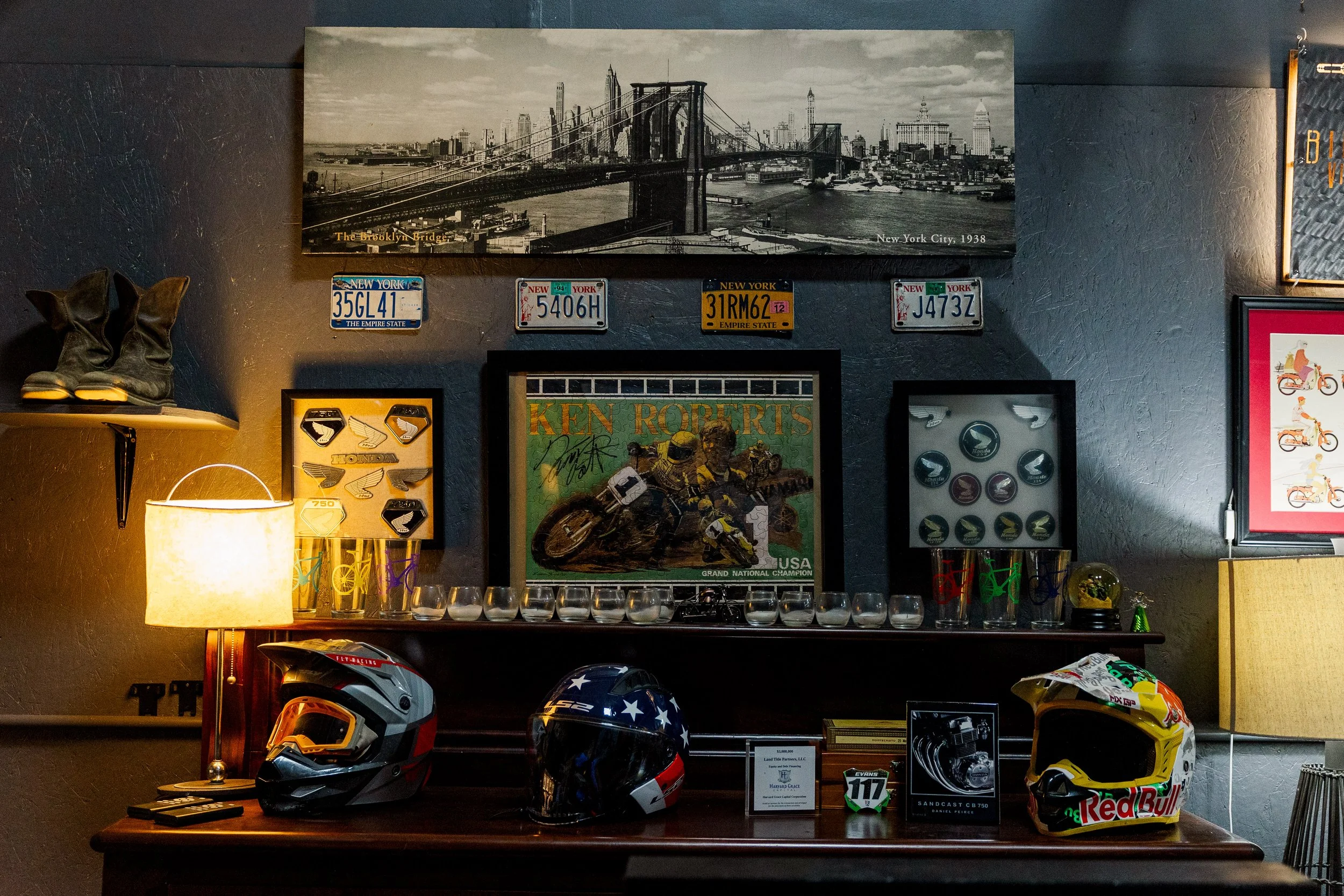 A wall decorated with framed pictures, vintage license plates, and motorcycle helmets, including a black helmet with stars and a yellow helmet with Red Bull branding. There is a Brooklyn Bridge poster and a framed picture of a motorcycle race on the 