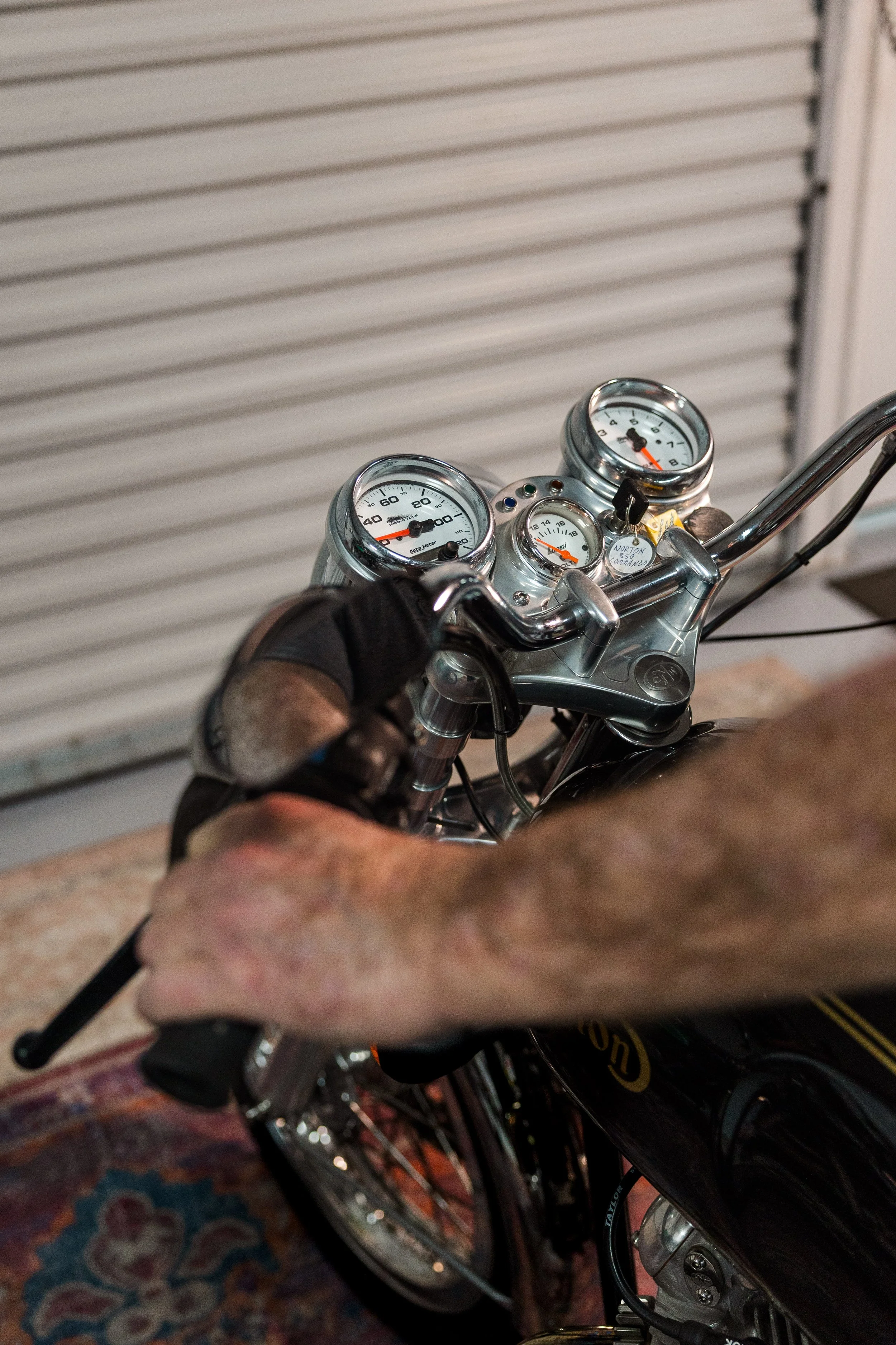 Close-up of motorcycle handlebars with a person's hand gripping the left handlebar, showing the speedometer and other gauges, and a background with a closed garage door. Vintage motorcycle detail at Birmingham Vintage Cycle, a vintage motorcycle rest