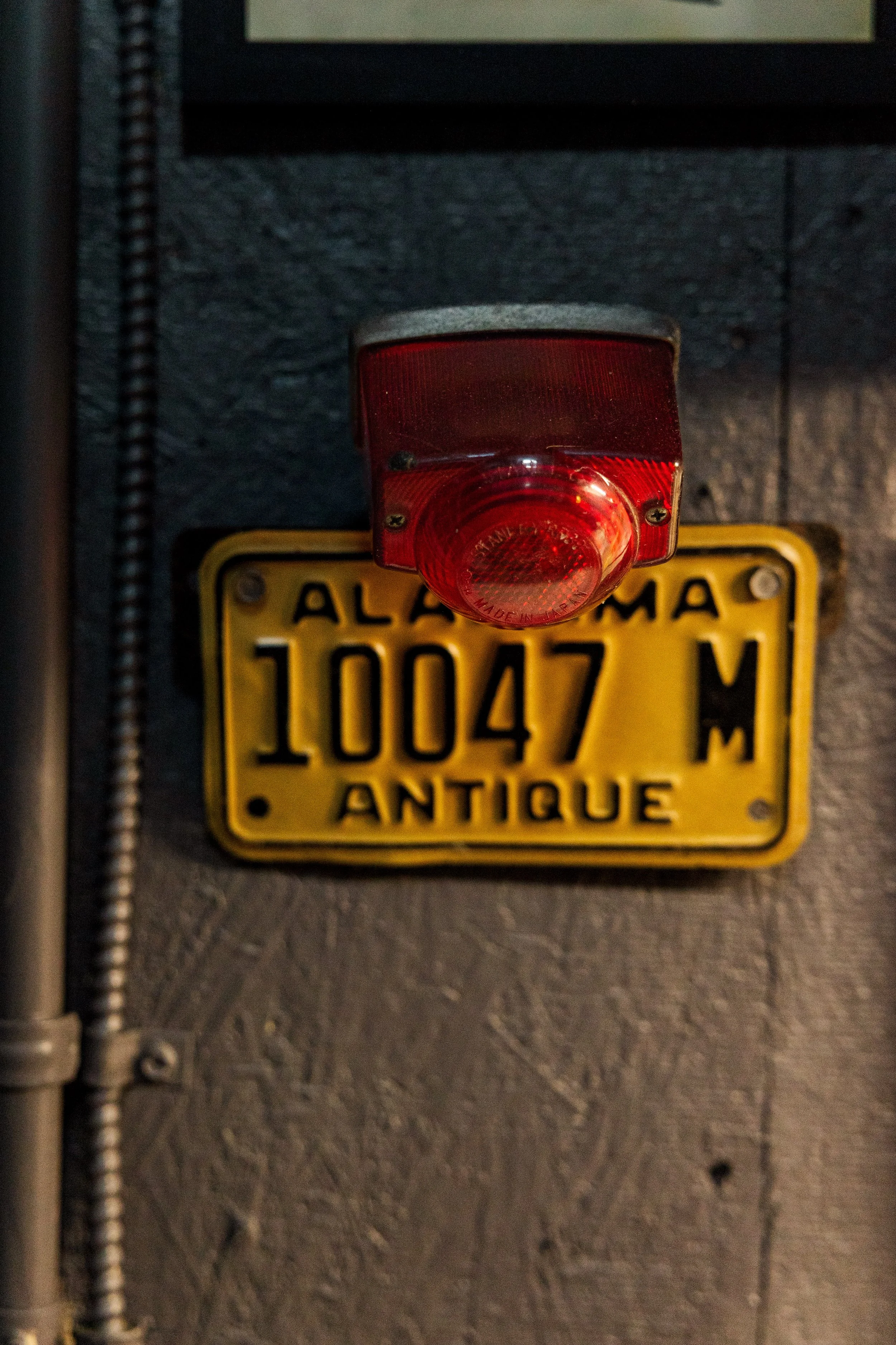A close-up of a yellow antique license plate from Alabama with the word 'ANTIQUE' at the bottom. Above it, there is a red reflector light mounted on a wall. The license plate number is 10047M. Part of an electrical conduit and a wire are visible on t