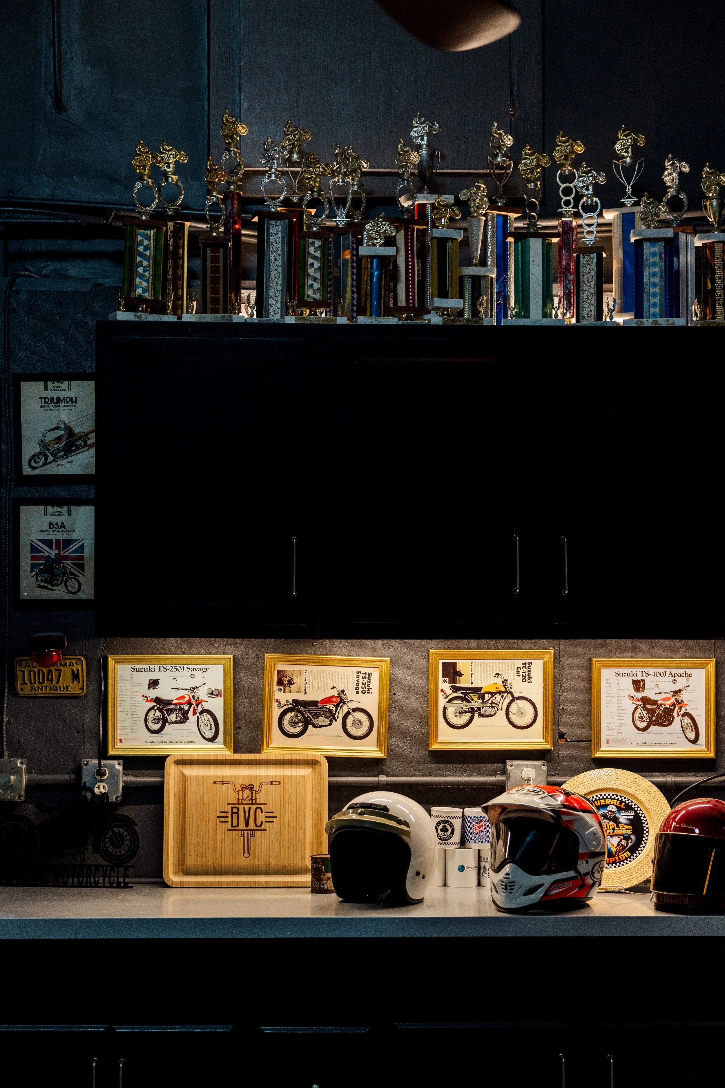 Display of motorcycle helmets and framed motorcycle posters on a dark wall in a motorcycle-themed shop or museum. Inside Birmingham Vintage Cycle, a vintage motorcycle shop and community space in Birmingham Alabama