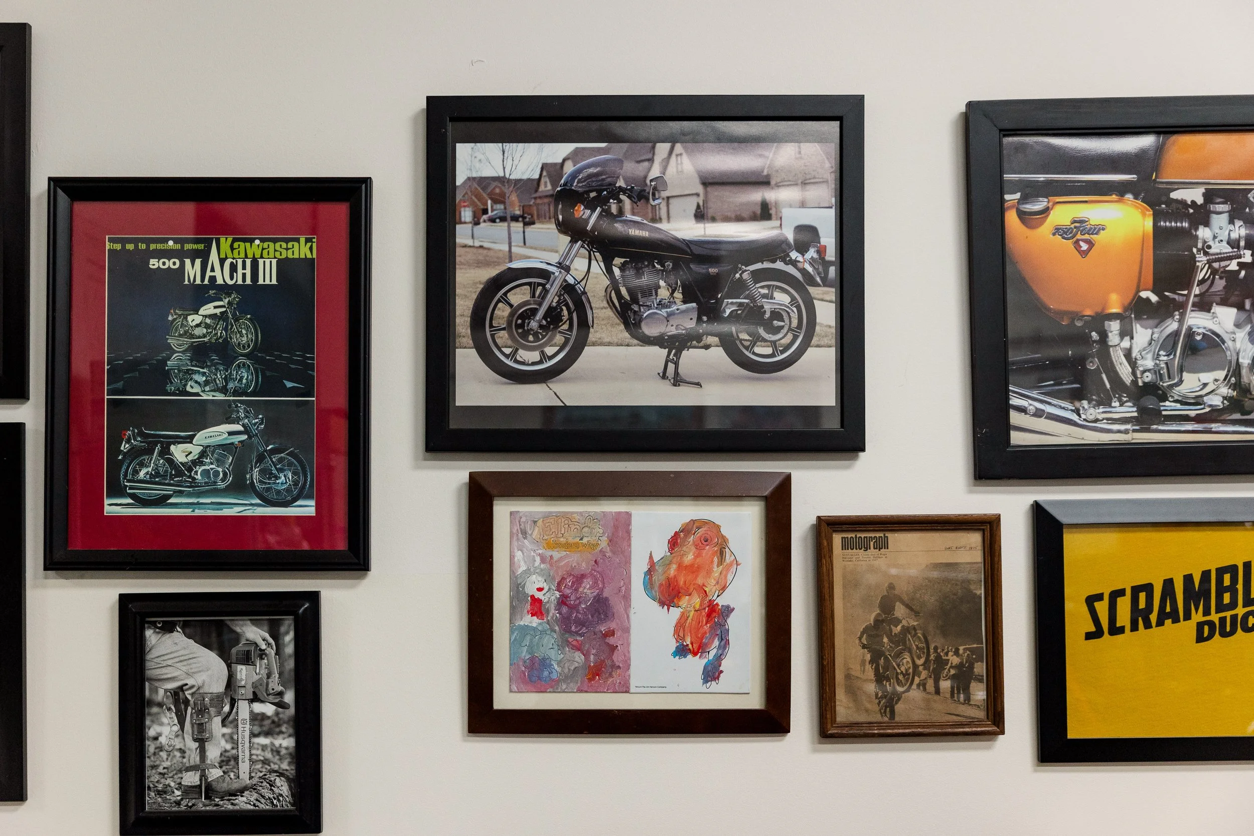 A collection of framed pictures and posters on a wall, featuring vintage motorcycles, artwork, and various illustrations. Inside Birmingham Vintage Cycle, a vintage motorcycle shop and community space in Birmingham Alabama