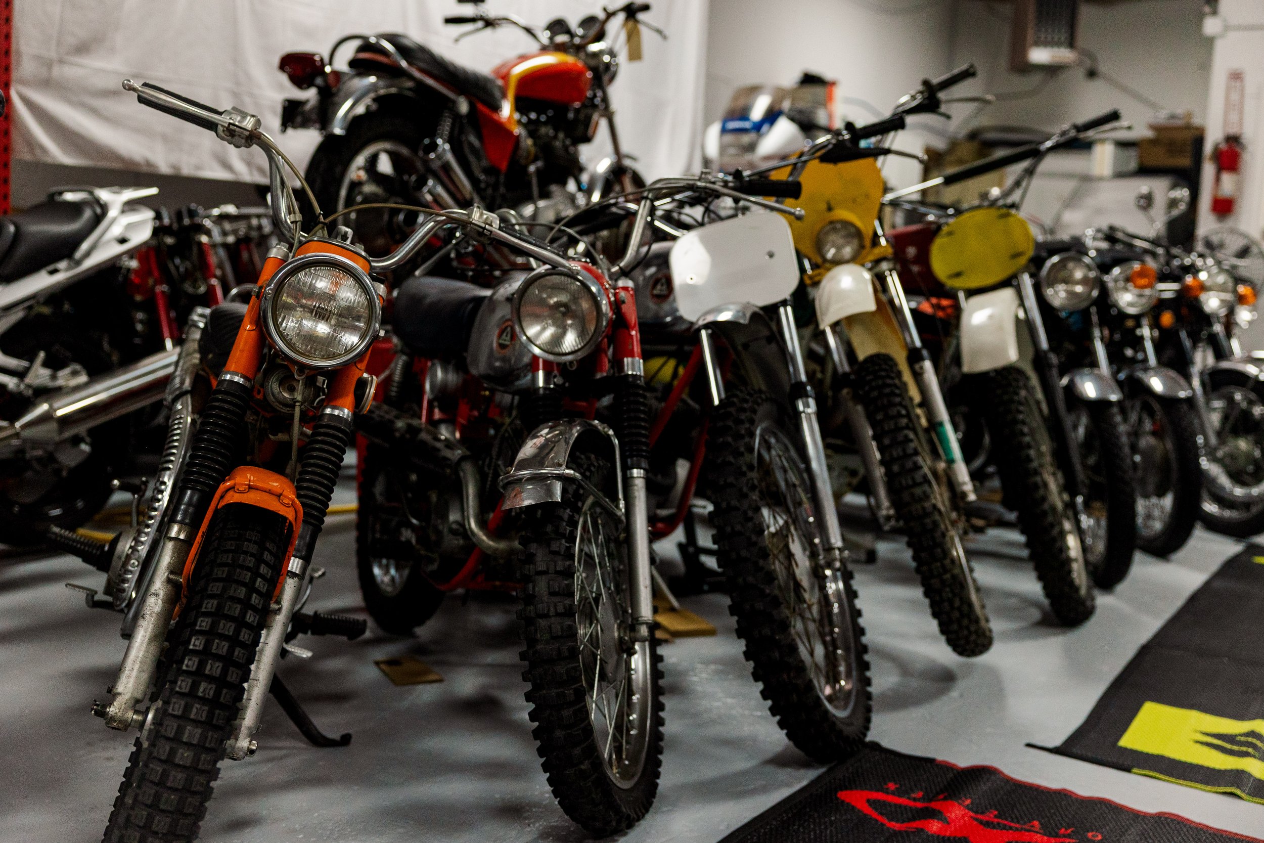 A row of vintage off-road motorcycles with knobby tires, parked indoors on a concrete floor. The motorcycles have various colors including orange, yellow, and white, and feature classic designs with round headlights and simple frames.