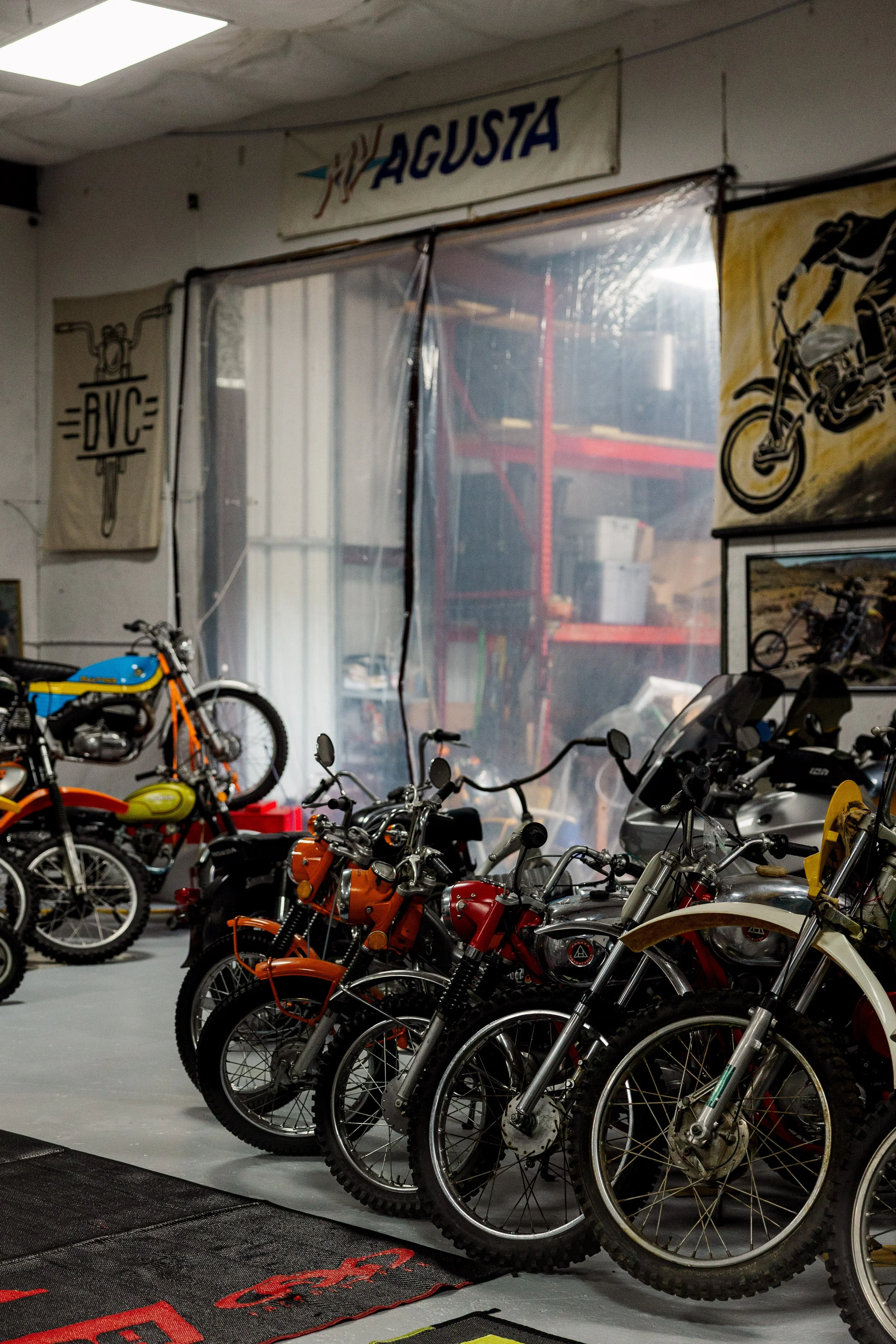 A collection of vintage motorcycles lined up inside a garage or showroom. The background features posters with motorcycle graphics and a sign that reads 'Agusta.'