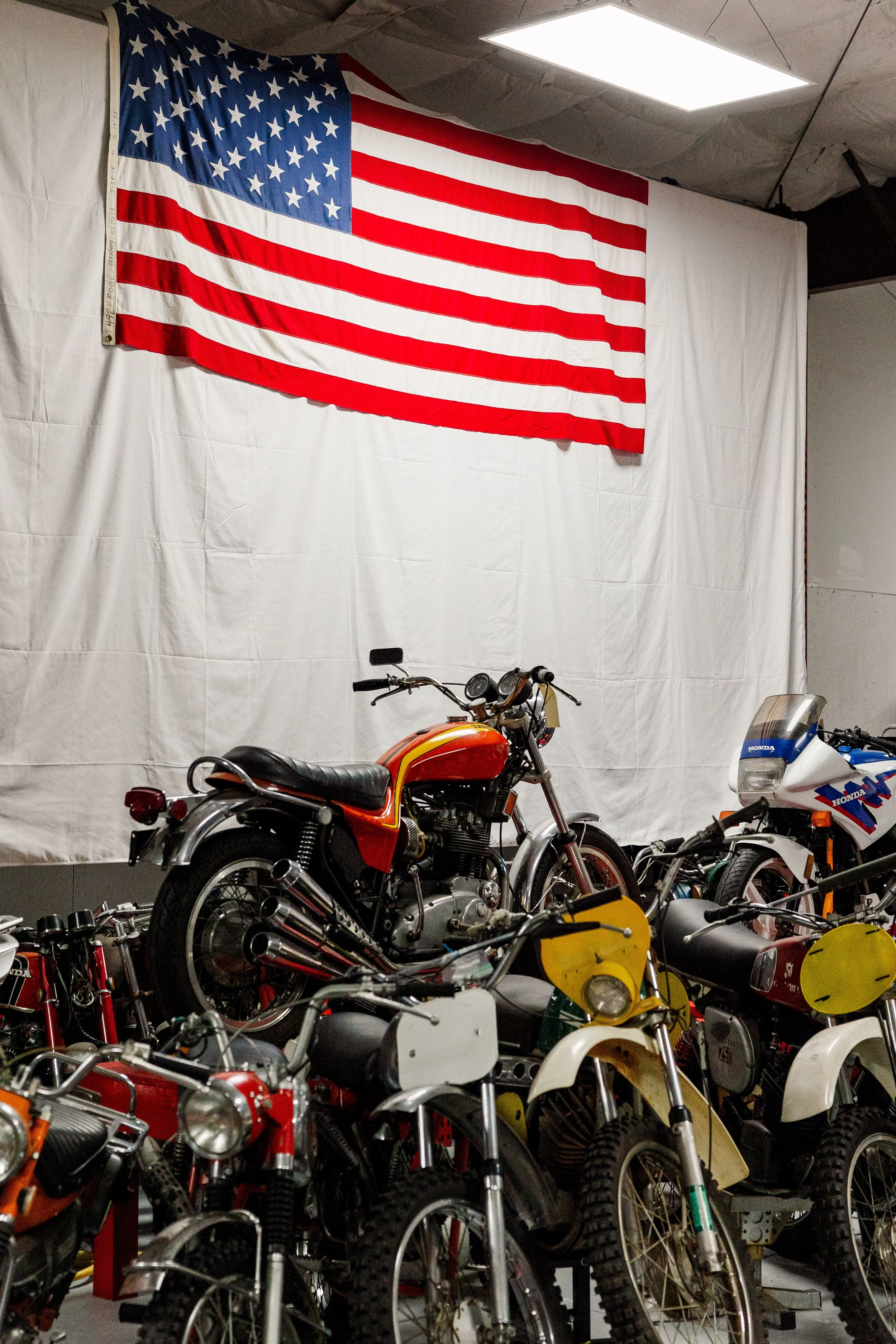 Multiple vintage motorcycles displayed in a shop with an American flag hanging on the wall in the background. Vintage motorcycle collection on display at Birmingham Vintage Cycle in Birmingham Alabama