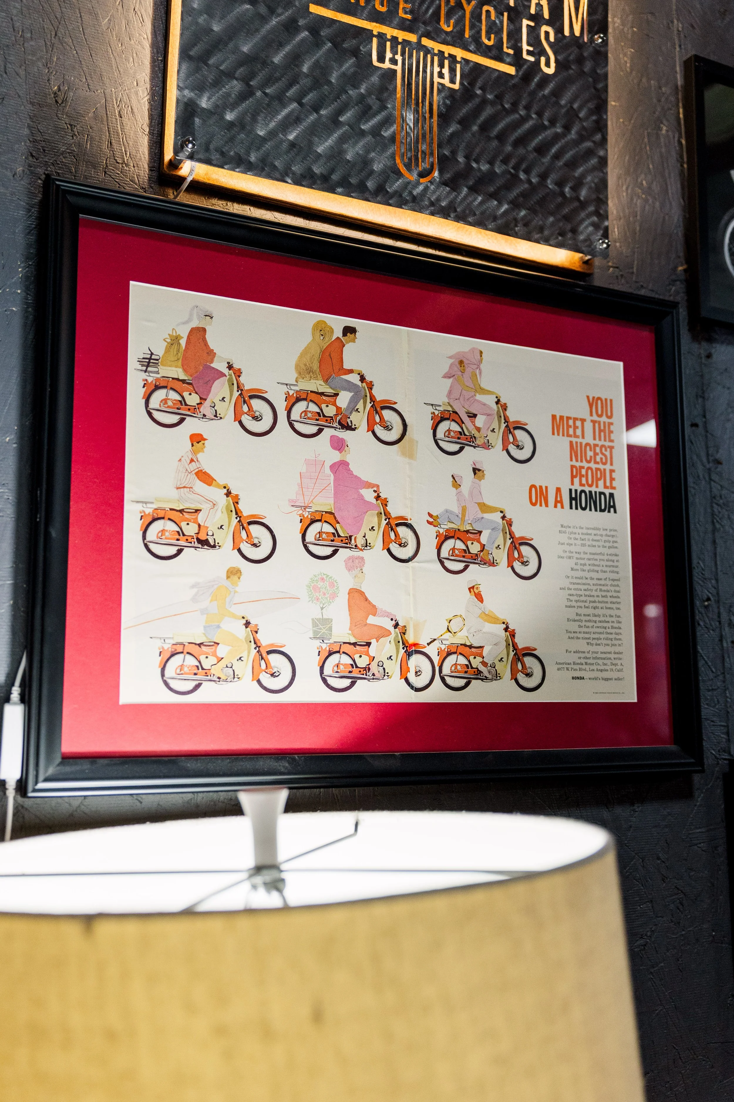Framed poster featuring illustrations of people riding orange Honda motorcycles, with a promotional slogan 'You meet the nicest people on a Honda' in orange text. Inside Birmingham Vintage Cycle, a vintage motorcycle shop and community space