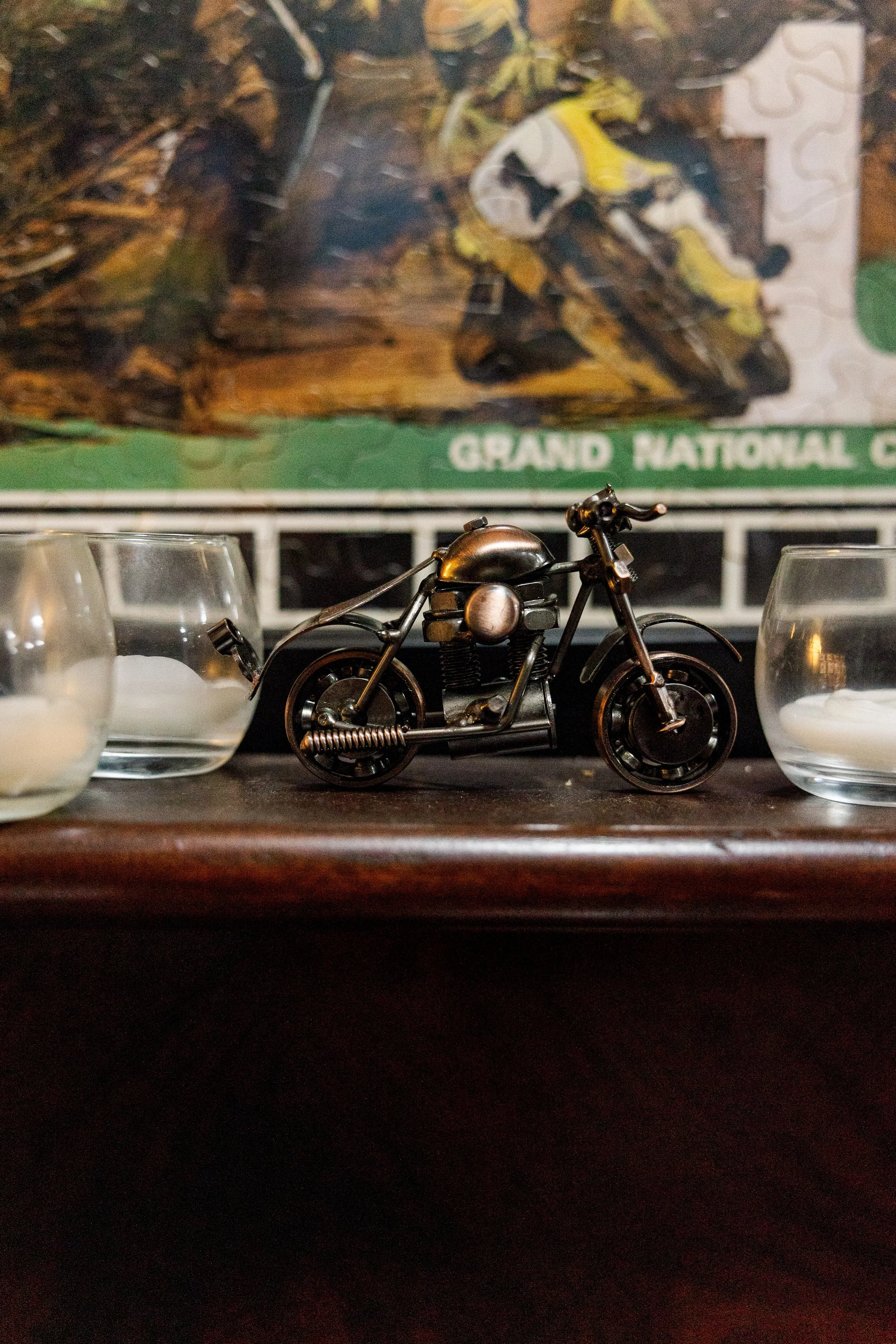 A small metallic model of a vintage motorcycle placed on a surface, with two clear glasses filled with a white beverage and a blurry background featuring a poster or picture with an outdoor scene. Inside Birmingham Vintage Cycle, a vintage motorcycle
