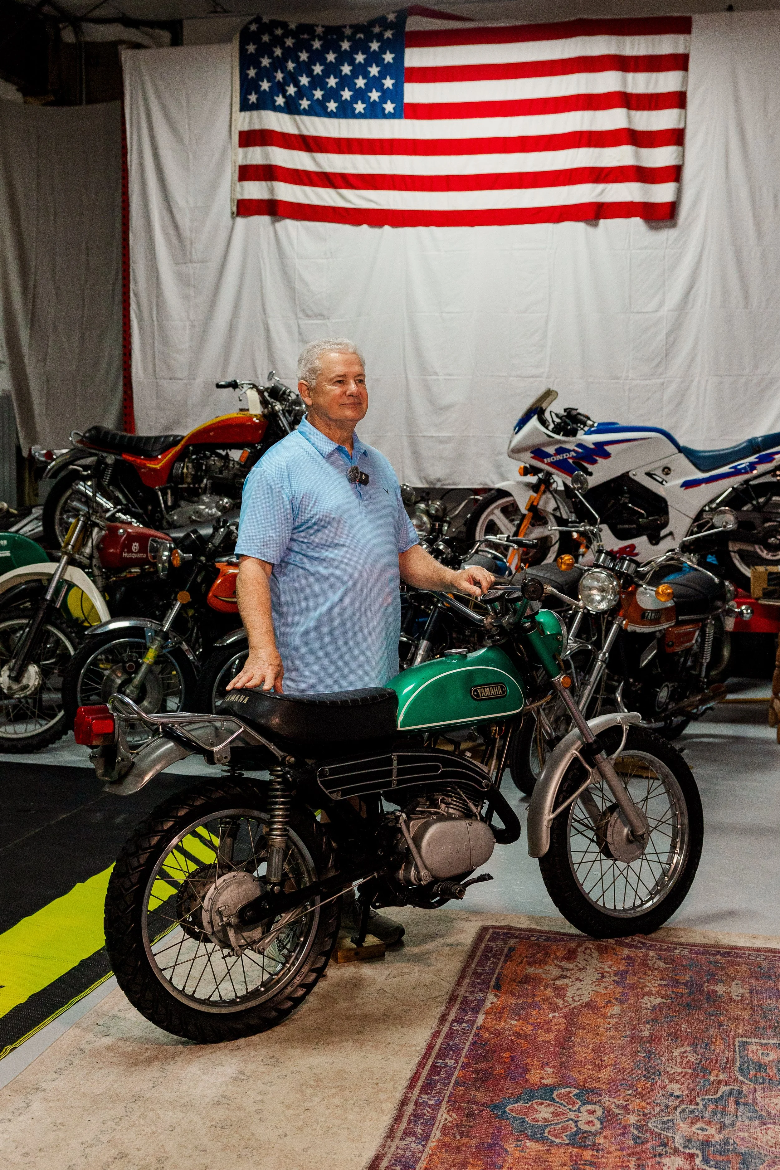 John Boone at BVC standing in a museum or showroom surrounded by vintage motorcycles. An American flag hangs on the wall behind him. The floor is partly covered with a patterned rug, and there is a yellow strip on the floor in the foreground.