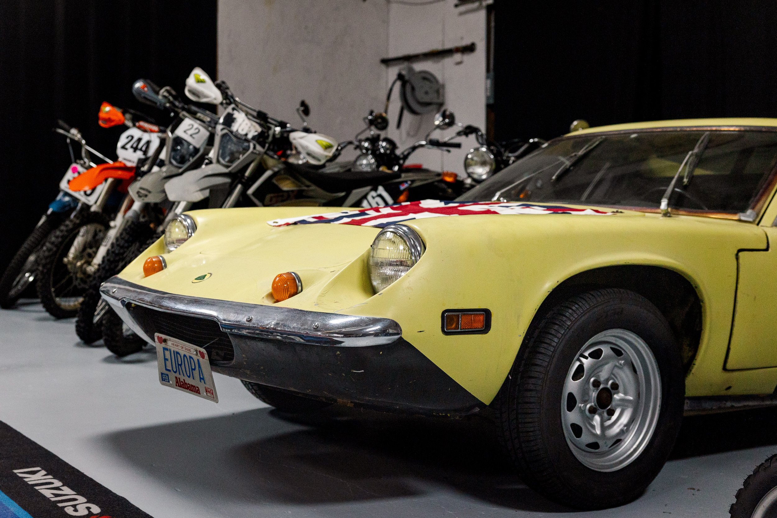 A vintage yellow sports car with an American flag decal on the hood, parked in a row with three white and black dirt bikes and a black motorcycle behind it in a dimly lit garage or showroom. Vintage motorcycle collection on display at BVC