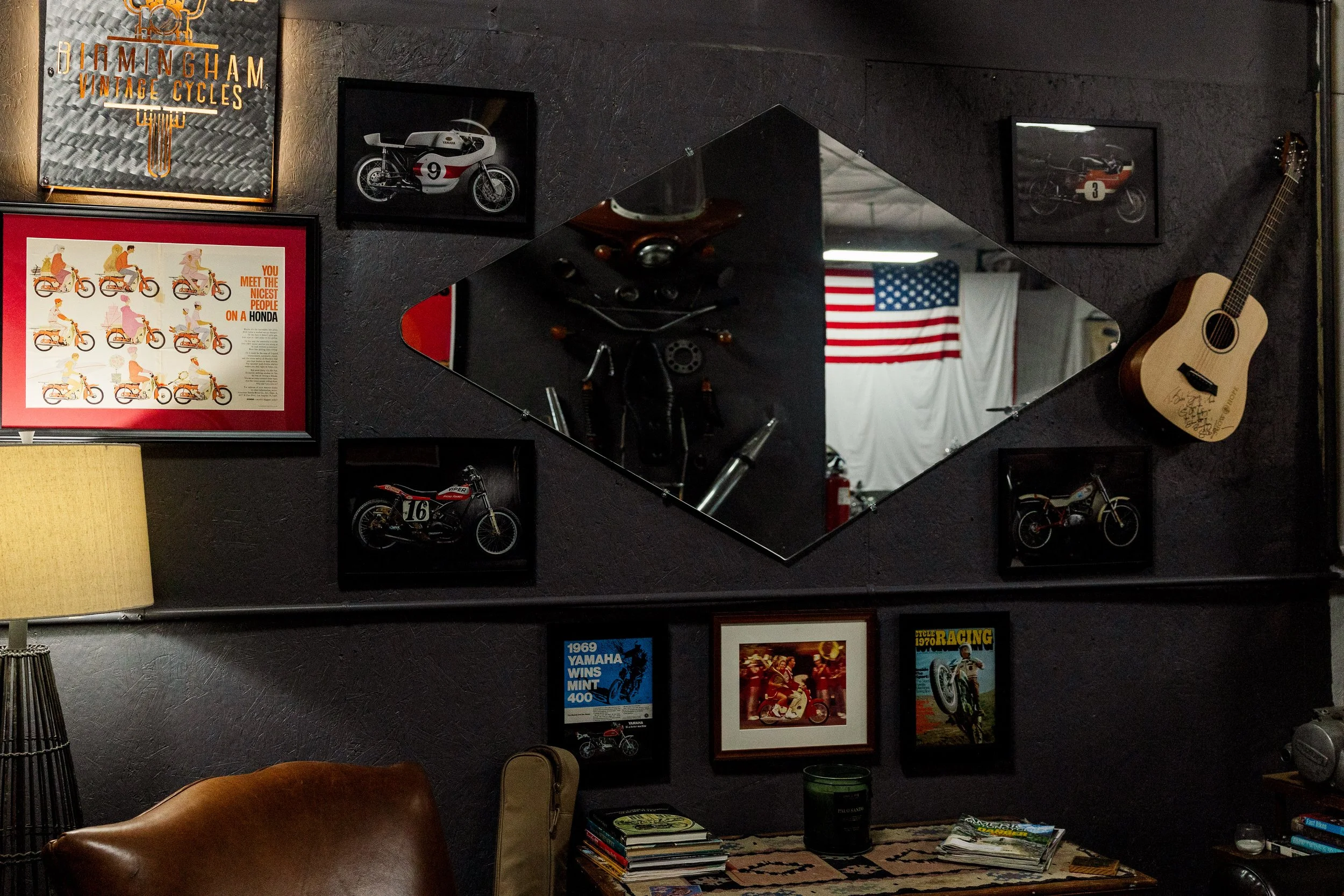 A wall decorated with vintage motorcycle and racing posters, photographs of motorcycles, and framed pictures. A guitar hangs on the right side of the wall. There is a large mirror reflecting part of the room, including an American flag, a fan, and va