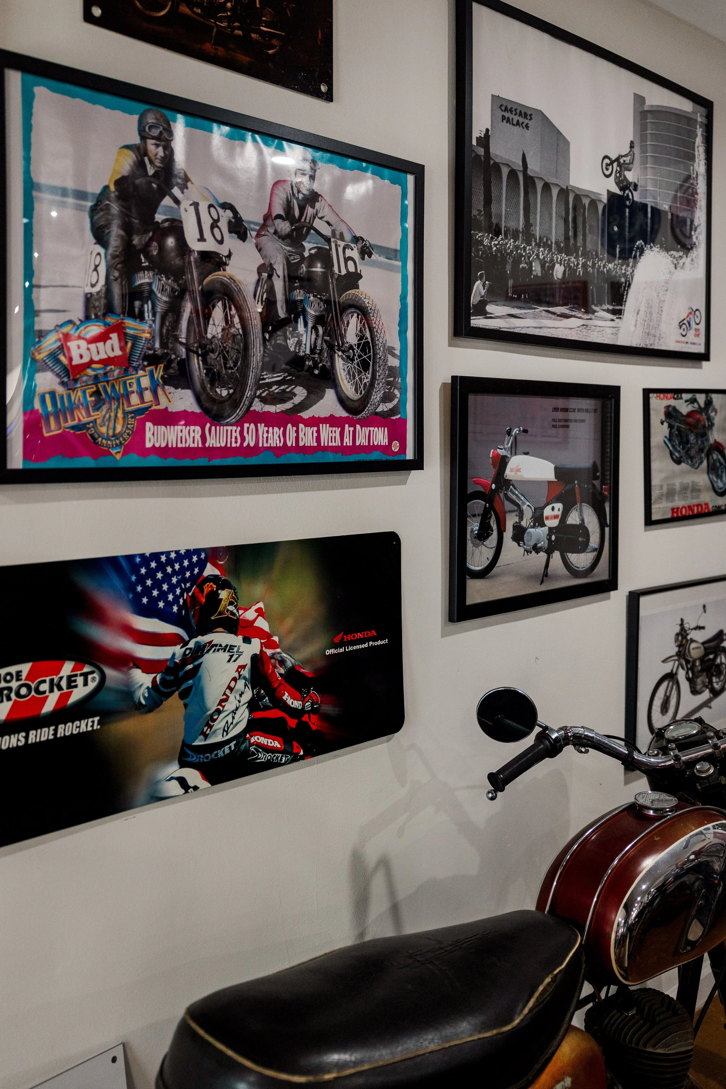 A collection of framed photographs on a wall, including a vintage motorcycle event poster, a black-and-white photo of Caesar's Palace, a vintage Honda motorcycle, and a motorcycle racer in action, with a motorcycle parked in front. Inside BVC