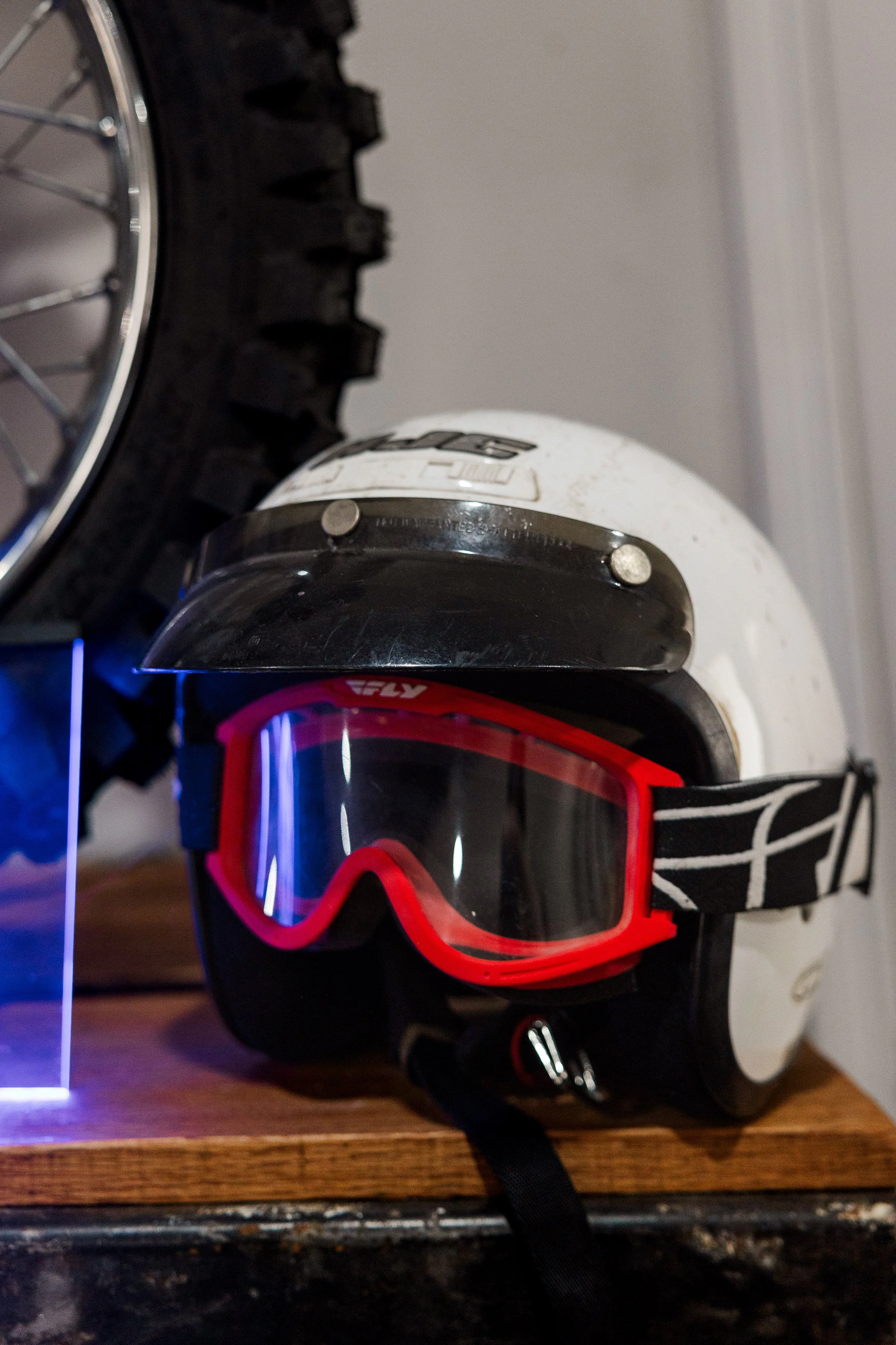 A pair of ski goggles with a red frame and black strap hanging on a hook next to a white ski helmet with black padding, set against a white wall. Inside Birmingham Vintage Cycle, a vintage motorcycle shop and community space in Birmingham Alabama