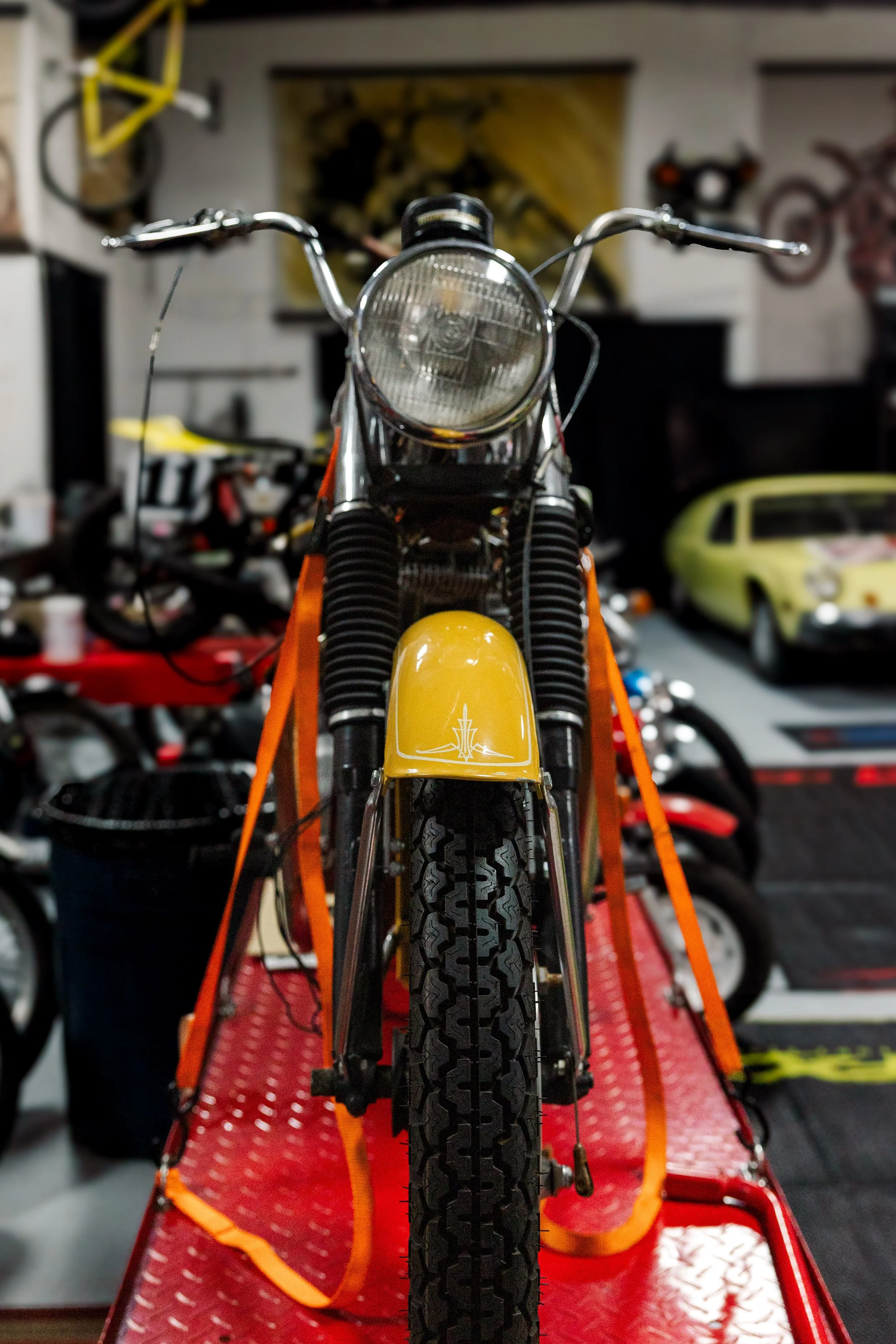 Front view of a vintage motorcycle with yellow front fender on a red platform, surrounded by various bikes and posters in a garage or workshop. Vintage motorcycle collection on display at Birmingham Vintage Cycle in Birmingham Alabama