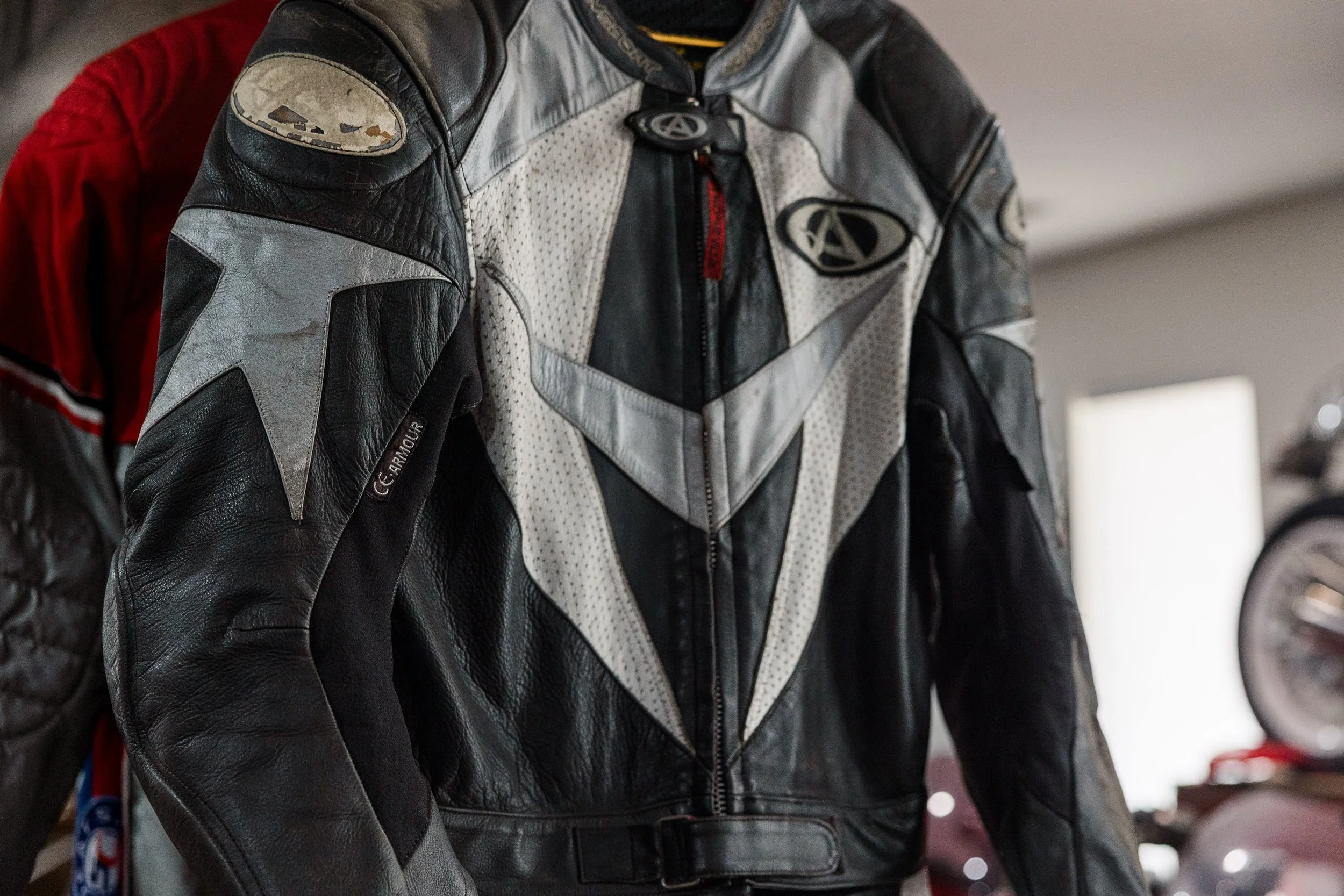 A motorcycle rider wearing a black and white leather riding jacket with patches and logos, standing indoors. Inside Birmingham Vintage Cycle, a vintage motorcycle shop and community space in Birmingham Alabama