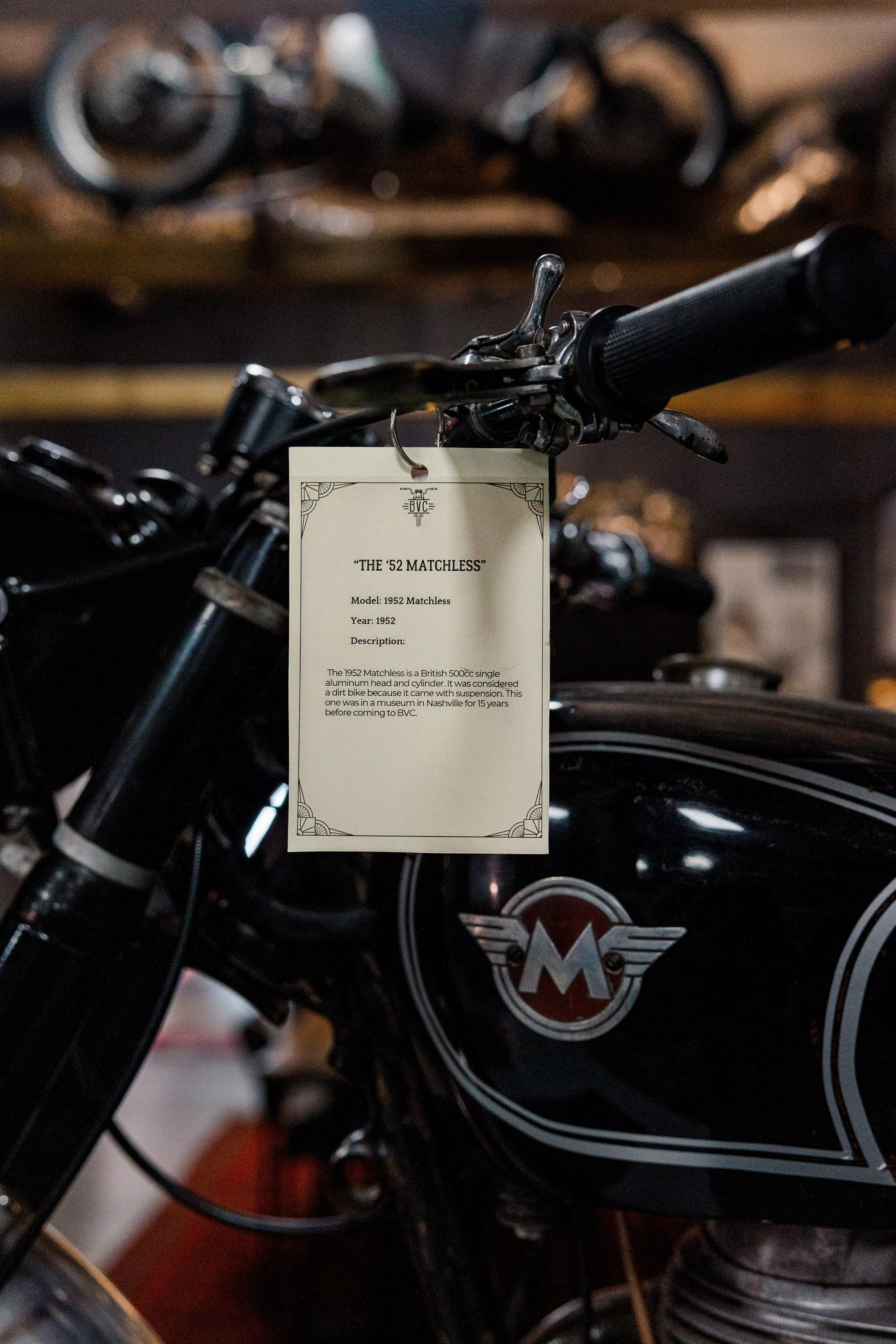 Close-up of a vintage motorcycle with a label that reads 'The '52 Matchless', displaying the model year 1952, parked among other motorcycles featuring a logo with a winged emblem. Vintage motorcycle collection on display at Birmingham Vintage Cycle 