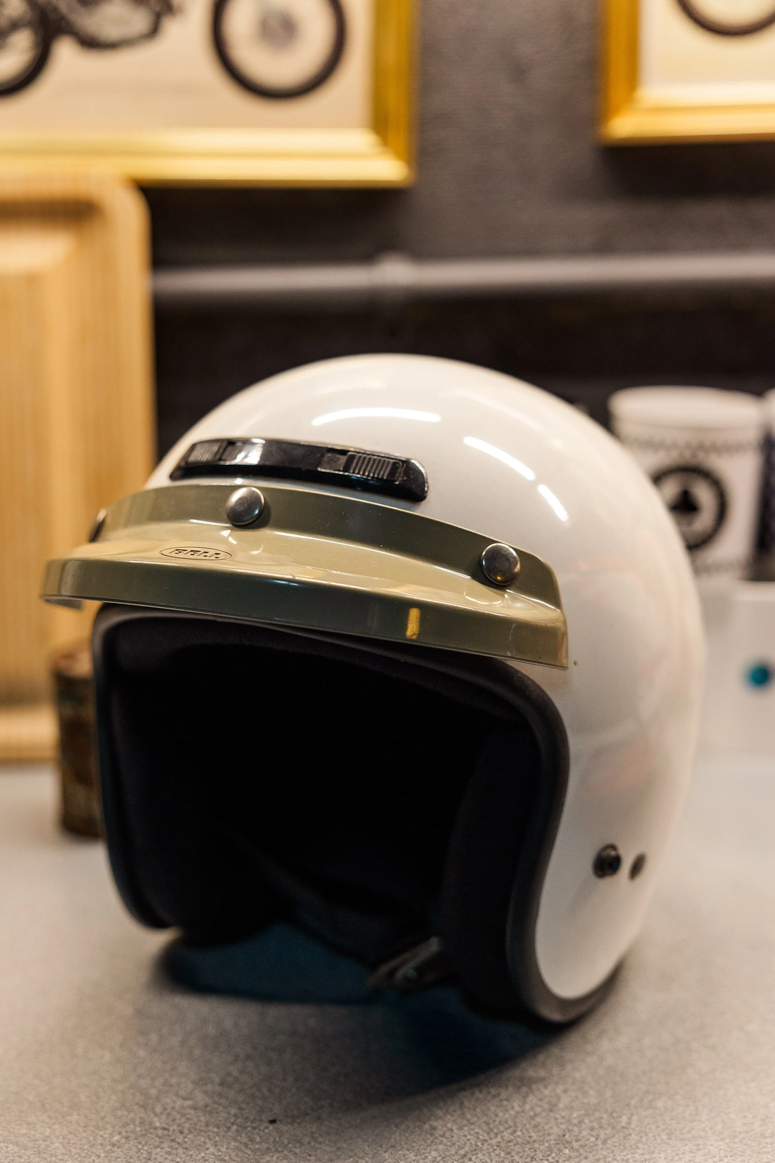 A white vintage-style electric helmet with black and silver accents placed on a gray surface, with a blurred background of framed artwork and a cup. Inside Birmingham Vintage Cycle, a vintage motorcycle shop and community space in Birmingham Alabama