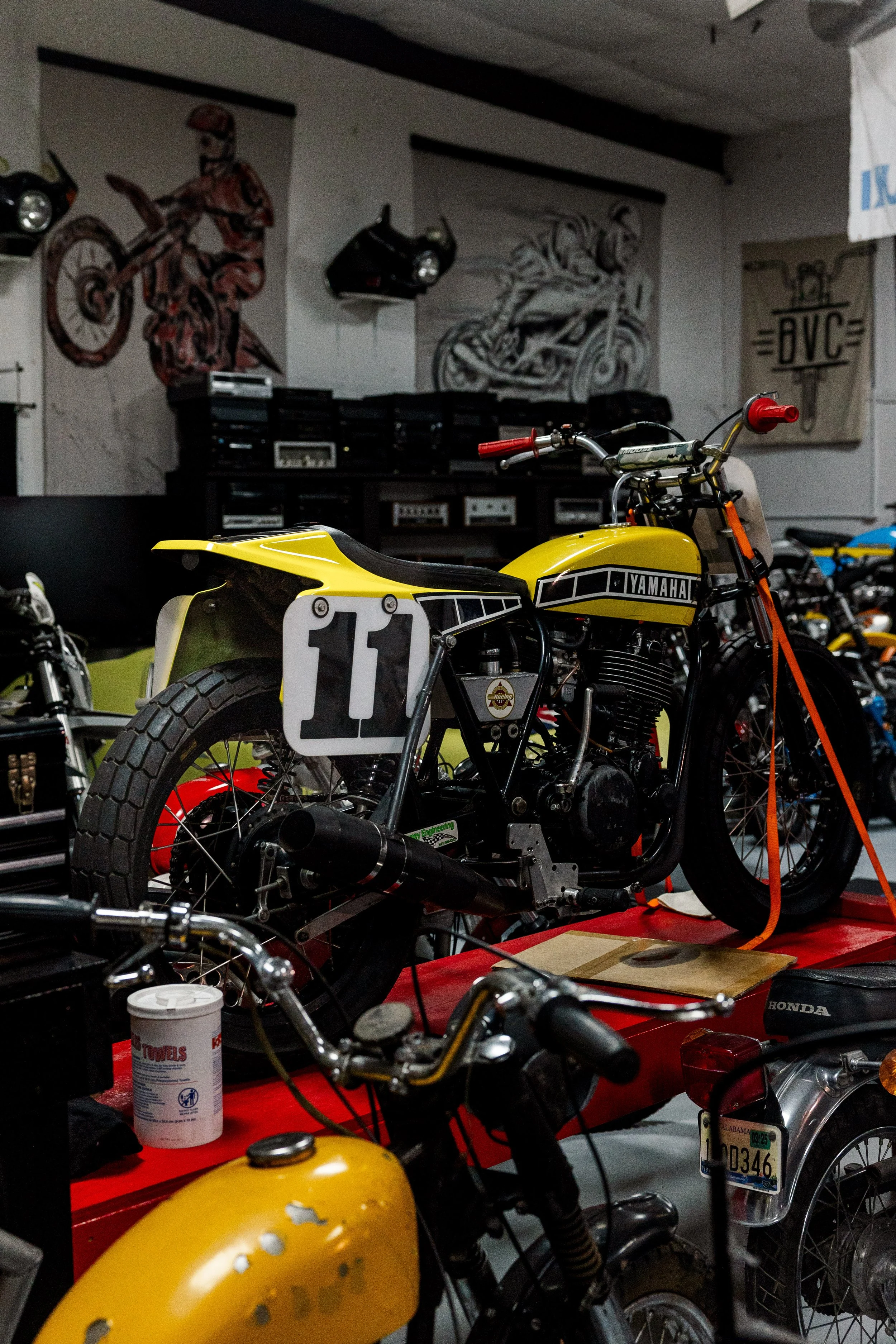 Indoor motorcycle garage with yellow Yamaha motorcycle in the foreground, other motorcycles, wall art, and equipment visible in the background. Vintage motorcycle collection on display at Birmingham Vintage Cycle in Birmingham Alabama
