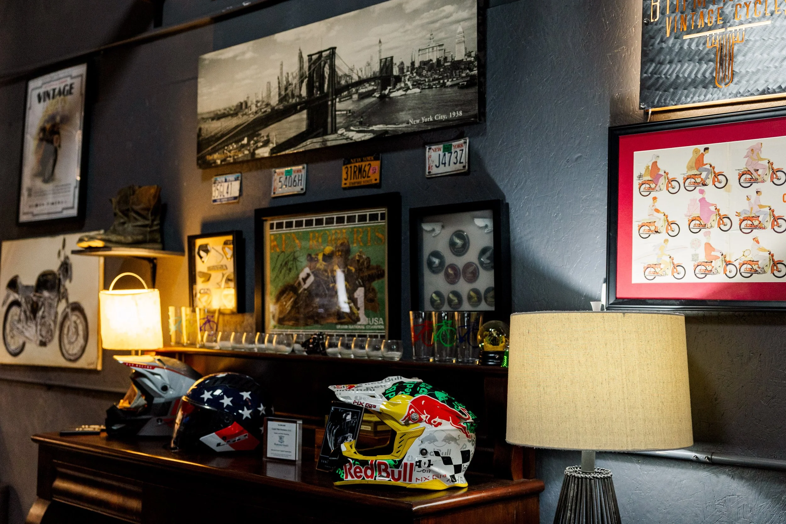 A collection of Brooklyn Nets and motorcycle helmets displayed on a wooden table. In the background, there are framed pictures and posters on a dark wall, including a black-and-white photograph of the Brooklyn Bridge and New York Cityscape from 1938,