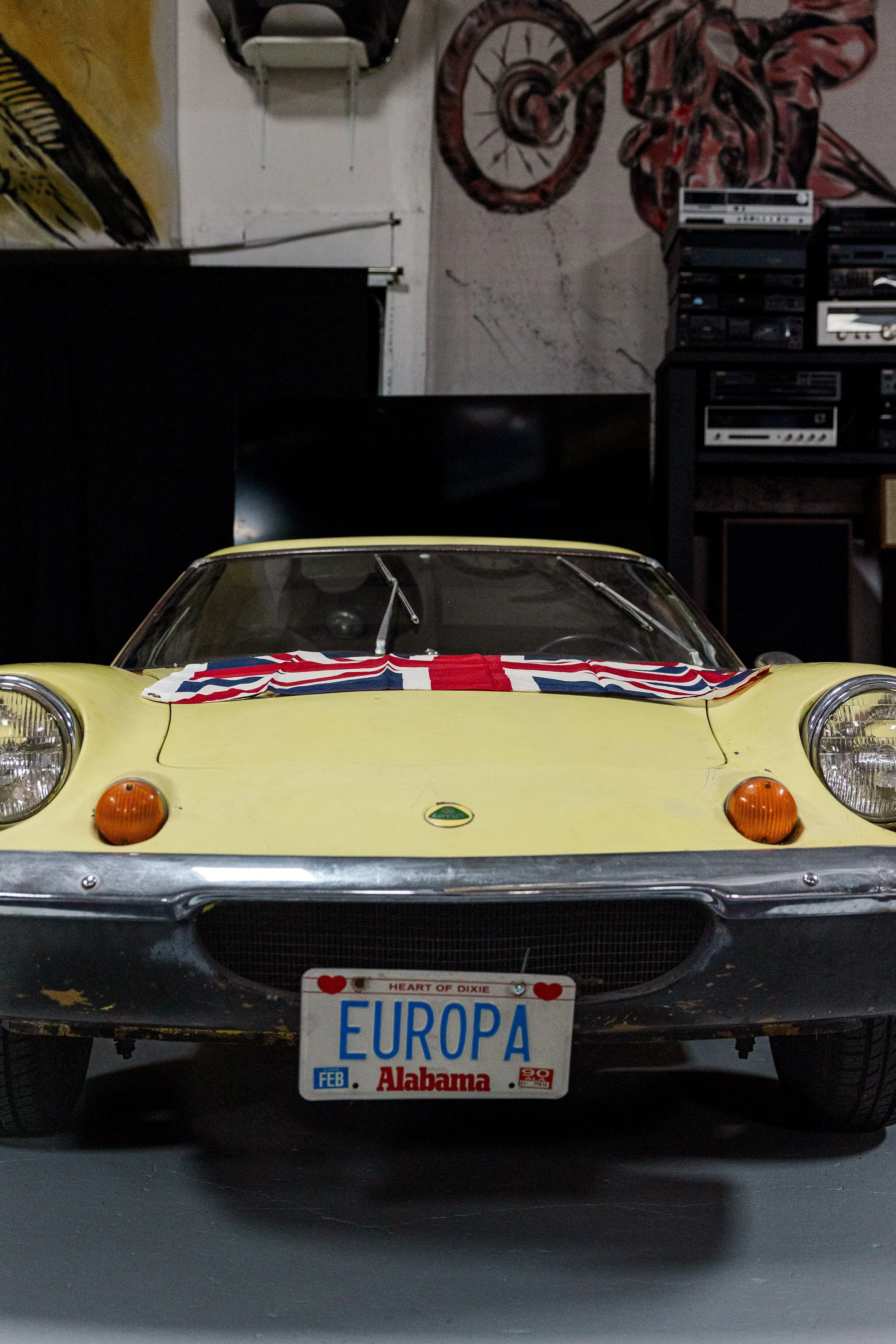 Front view of a vintage yellow Lotus sports car with a British flag on the hood, parked indoors, with an Alabama license plate reading 'EUROPA' and 'HEART OF DIXIE' at the front. The background features a wall with a motorcycle painted on it, a stere