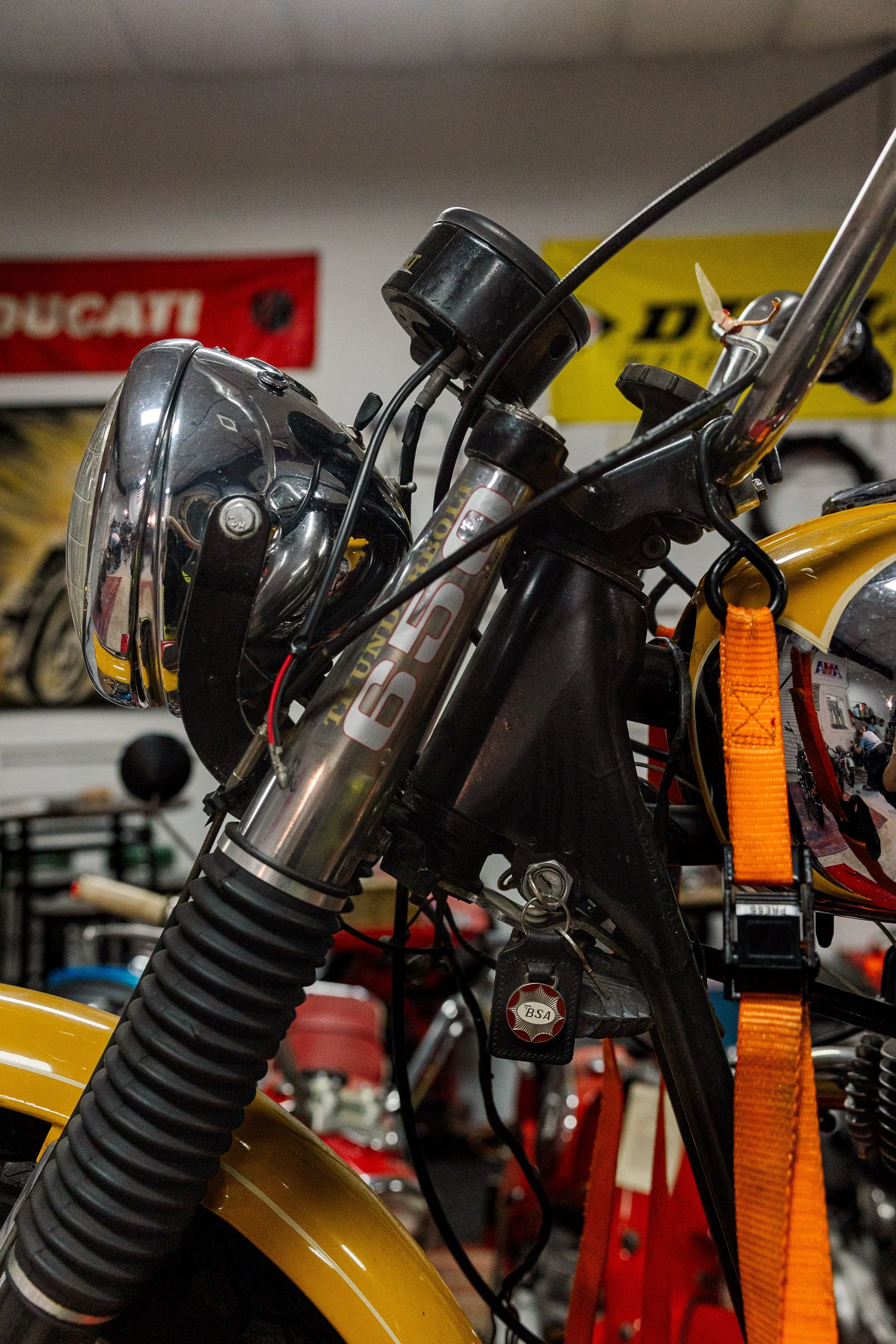 Close-up of a motorcycle's front section, showing the headlight, front suspension, and handlebars, with tools and equipment in the background. Vintage motorcycle collection on display at Birmingham Vintage Cycle in Birmingham Alabama