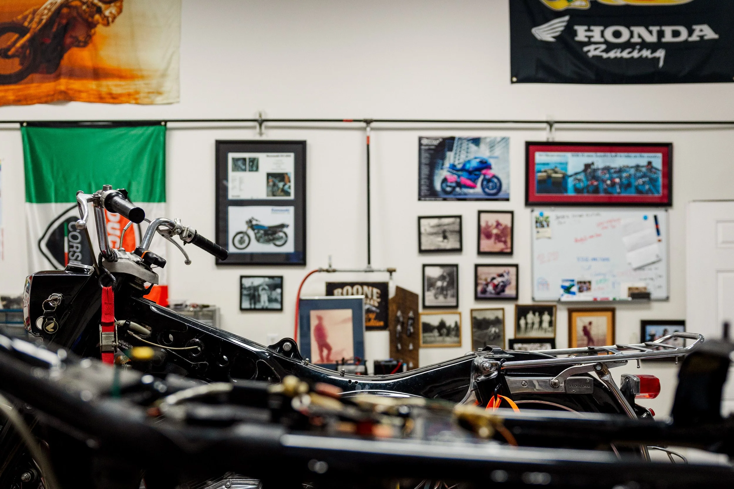 Partially assembled motorcycle in workshop with various framed pictures on the wall, including a motorcycle poster and Honda Racing banner. Inside Birmingham Vintage Cycle, a vintage motorcycle shop and community space in Birmingham Alabama