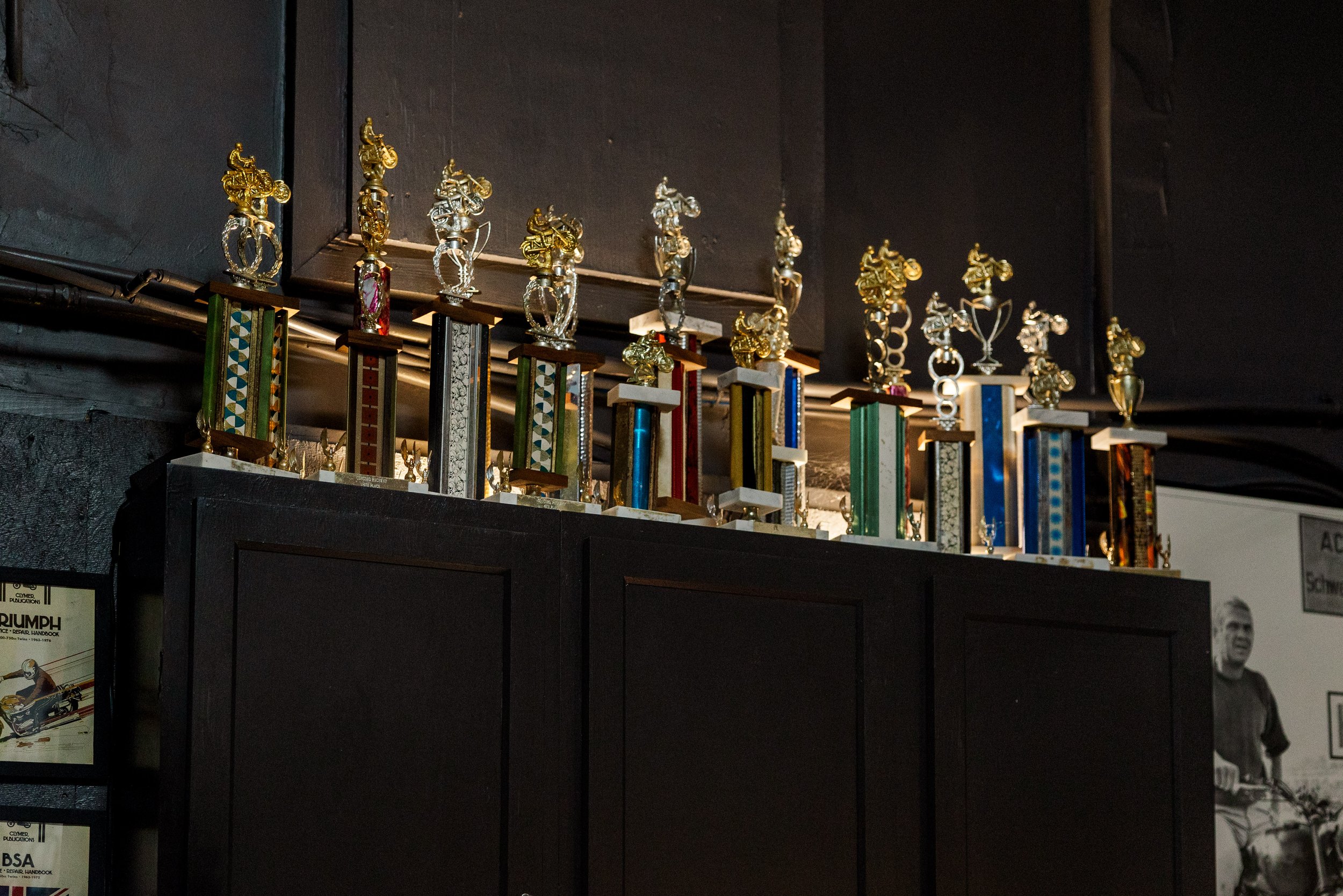 A collection of trophies with motorcycle rider figures on top, arranged on a dark shelf against a dark wall. Inside Birmingham Vintage Cycle, a vintage motorcycle shop and community space in Birmingham Alabama