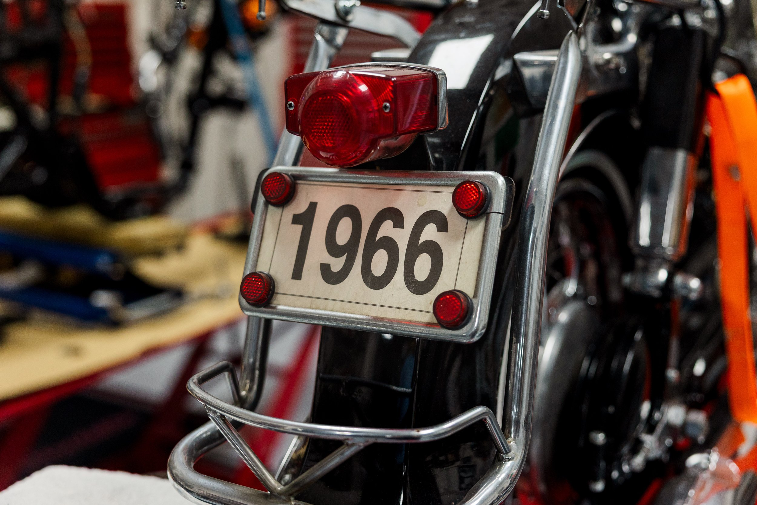 Close-up of a motorcycle rear end featuring a red tail light, license plate number 1966, and reflectors, with other motorcycles blurred in the background. Vintage motorcycle detail at Birmingham Vintage Cycle, a vintage motorcycle restoration shop in