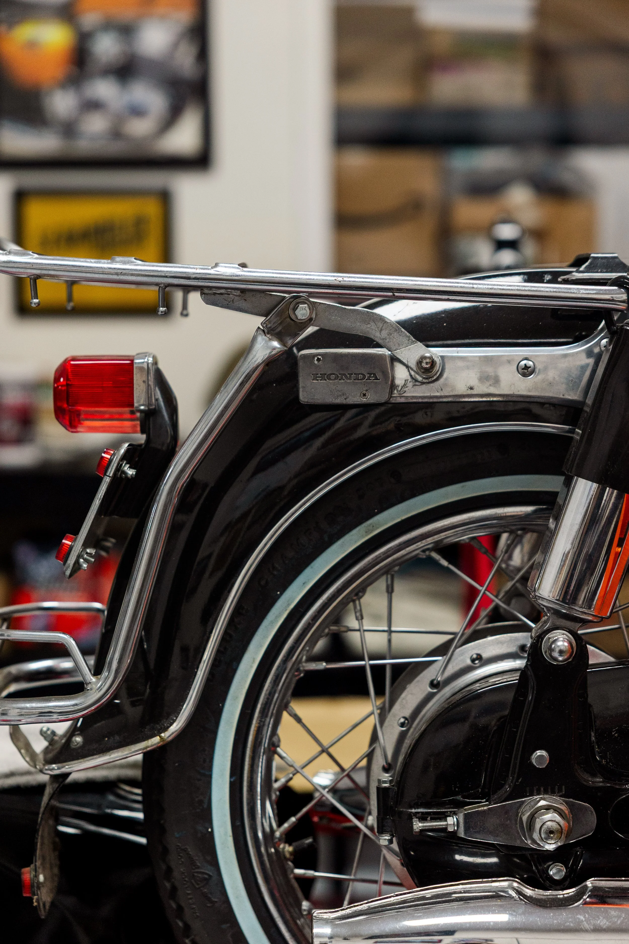 Close-up of a chrome wheelchair with a black tire, red reflectors, and various mechanical parts, in a workshop or storage room with shelves and posters in the background. Vintage motorcycle detail at Birmingham Vintage Cycle, a vintage motorcycle 