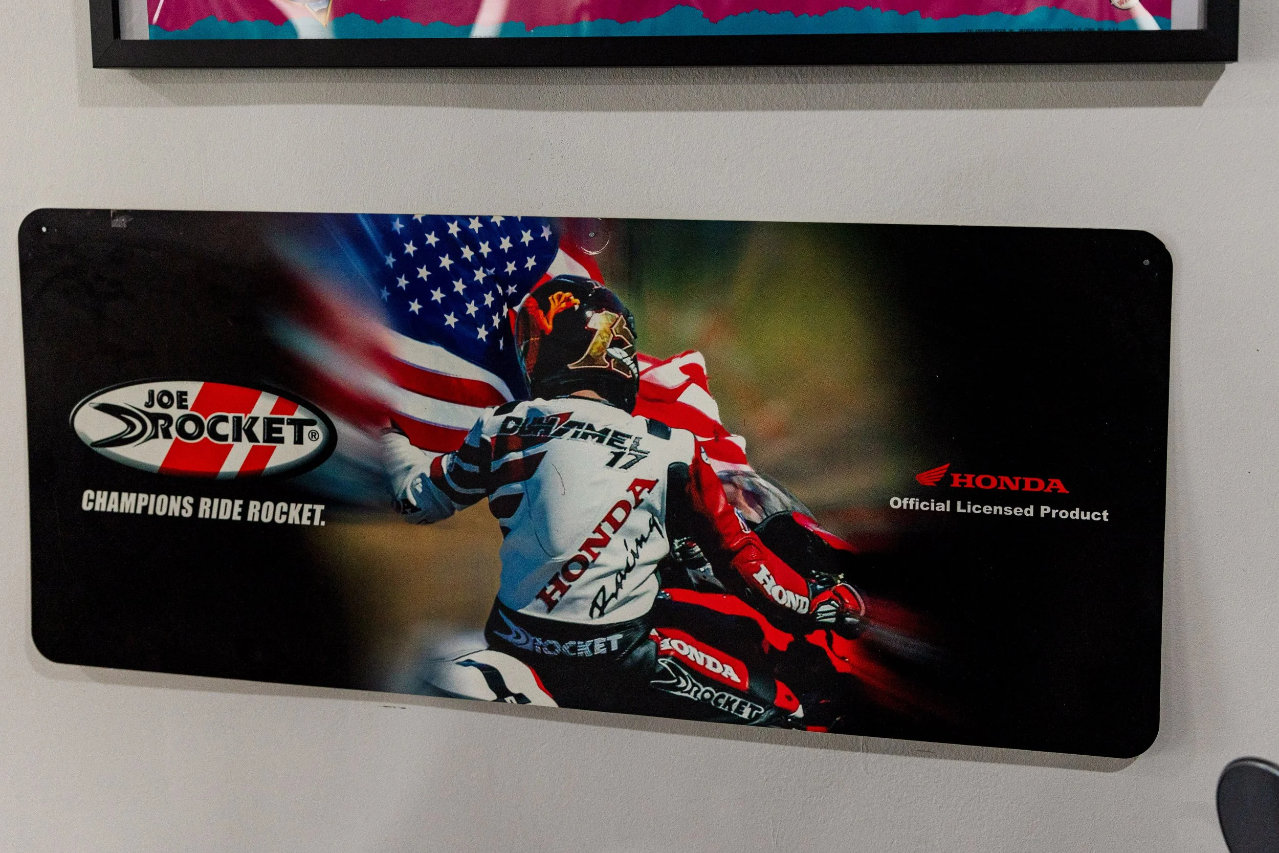 Poster of a motorcycle race featuring a rider in red, white, and blue gear, holding a helmet, with an American flag background and 'Joe Rocket' and 'Honda' logos. Vintage motorcycle collection on display at Birmingham Vintage Cycle in Birmingham