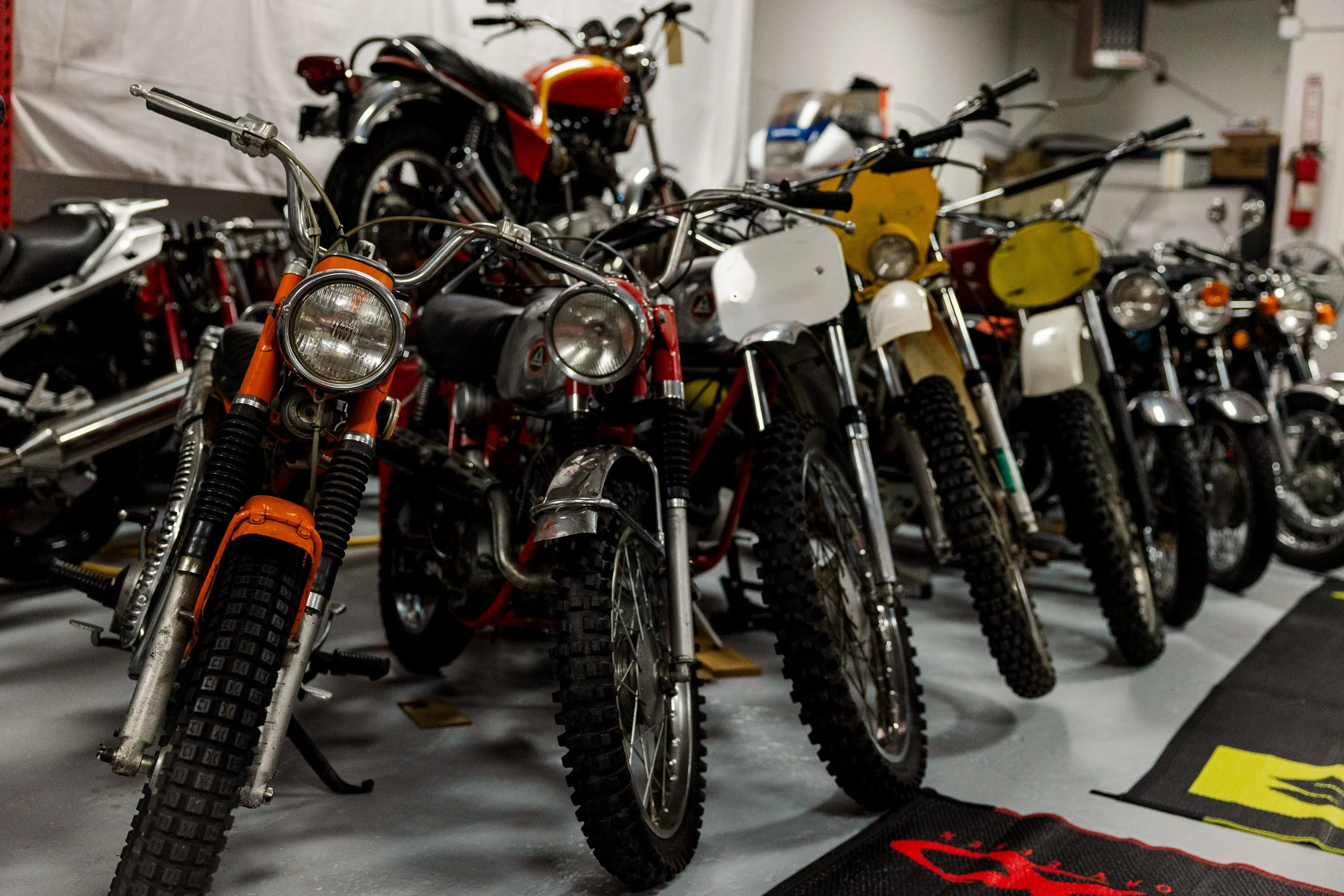 A row of vintage dirt bikes with knobby tires and various colors parked indoors, with some motorcycles stacked and stored in the background. Vintage motorcycle collection on display at Birmingham Vintage Cycle in Birmingham Alabama