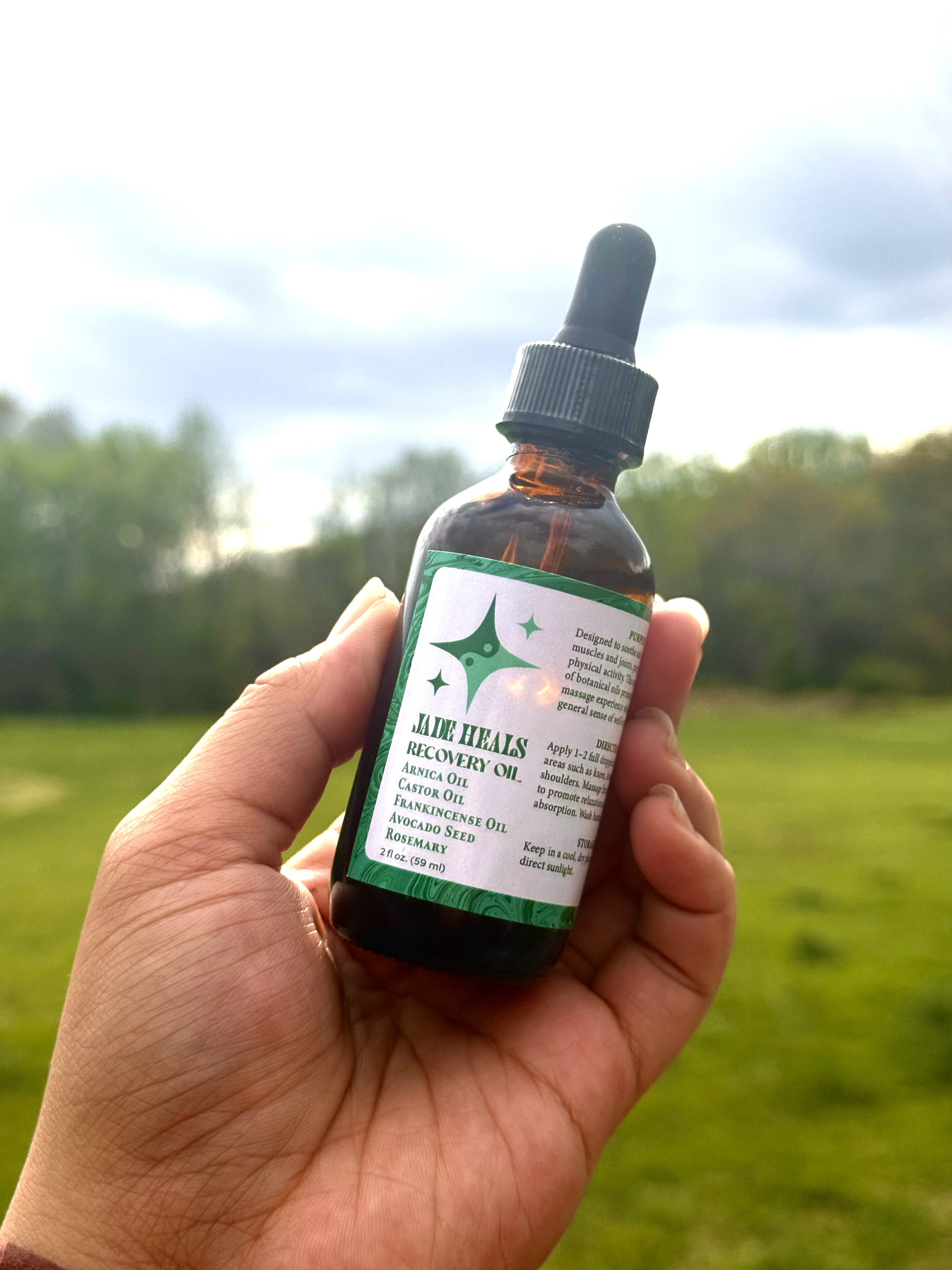 A person holding a small amber glass bottle with a black dropper cap outdoors against a blurred background of green grass and trees. The bottle's label reads "Jade Heals Recovery Oil," listing ingredients such as Arnica Oil, Castor Oil, Frankincense Oil, Avocado Seed, and Rosemary.