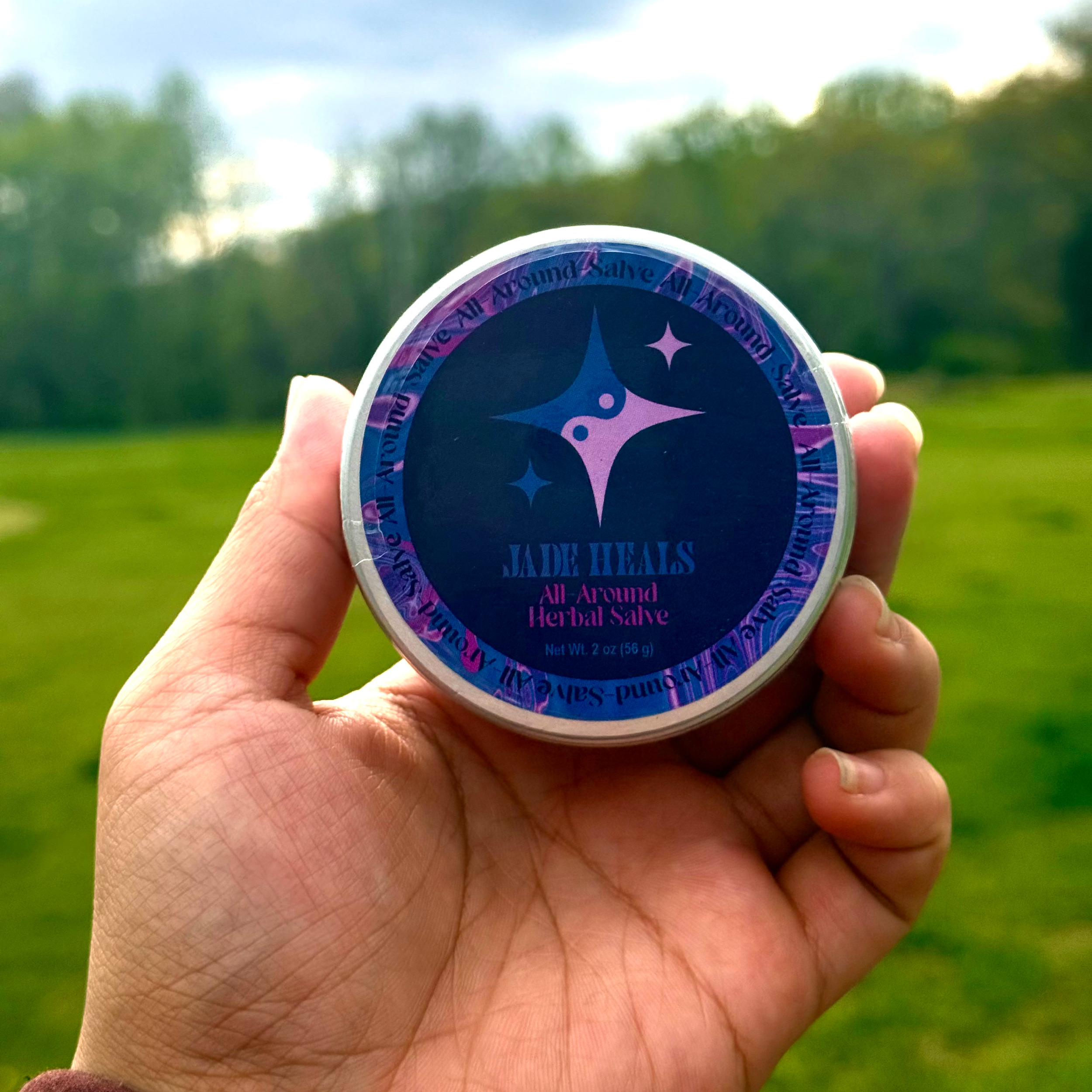 A hand holding a small container with a herbal salve label in front of a blurred outdoor background of trees and sky.