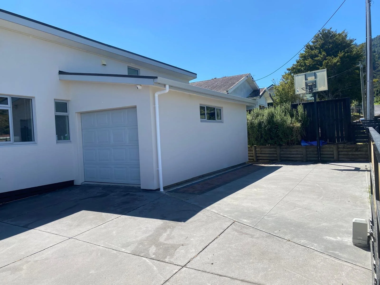 Backyard with a concrete patio, white house with garage door, basketball hoop, and wooden fence, with hills and trees in the background.