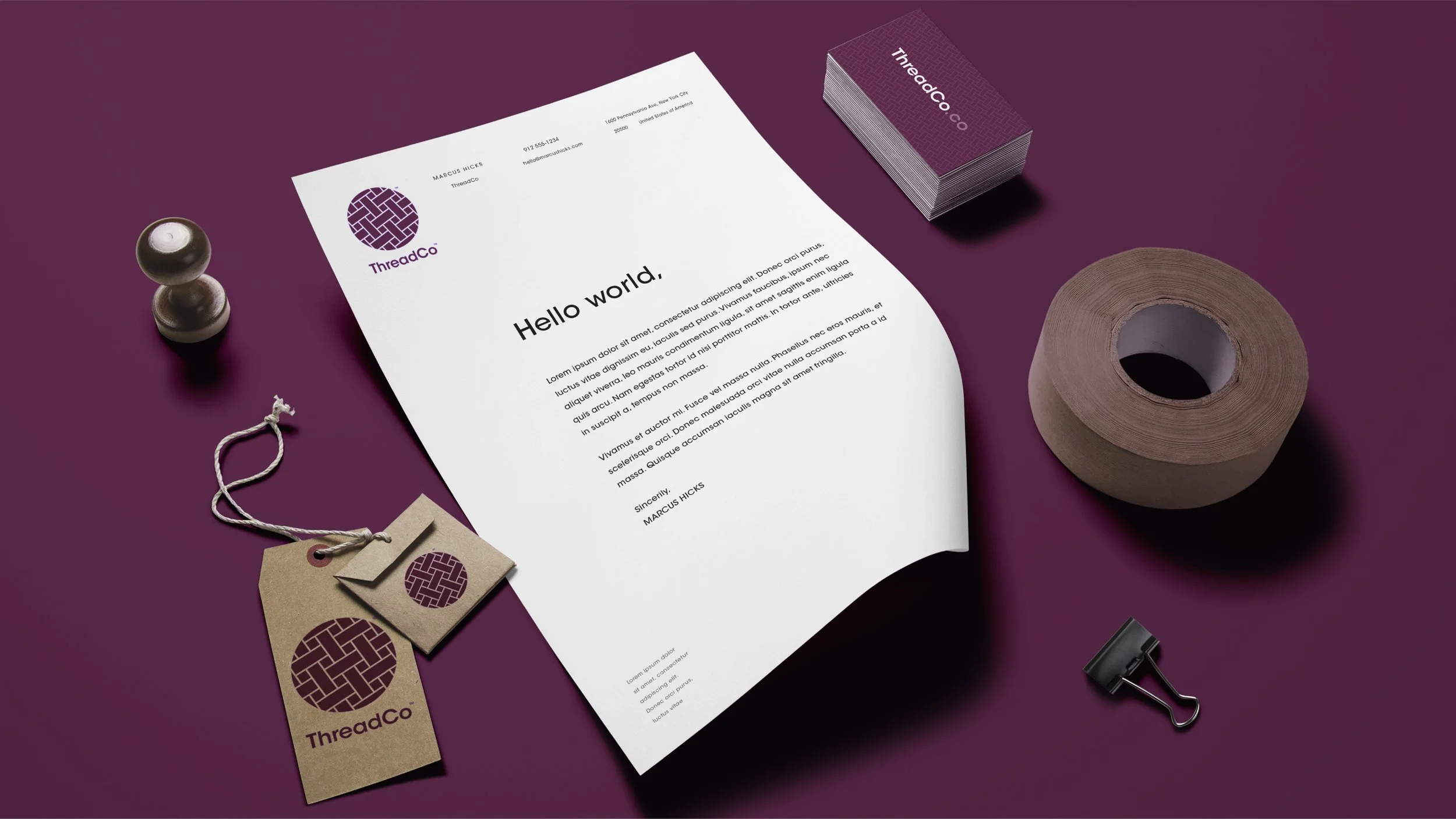 A purple desk with branded stationery items including a letter with the headline 'Hello world,' business cards, tags, a roll of paper, a black binder clip, and a small figurine.