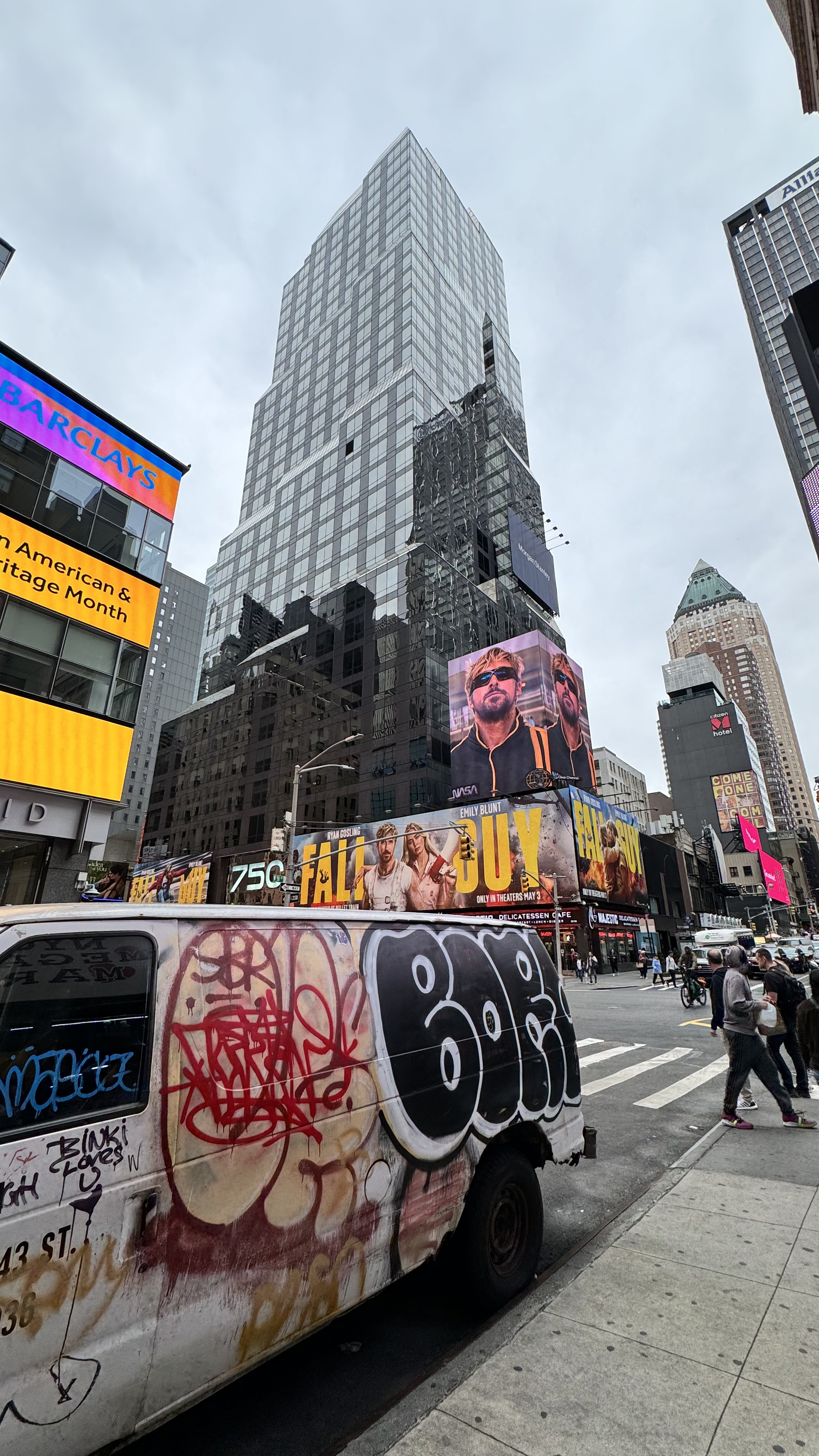 City scene with tall skyscraper, digital billboards, busy street, graffiti-covered van, pedestrians, and overcast sky.